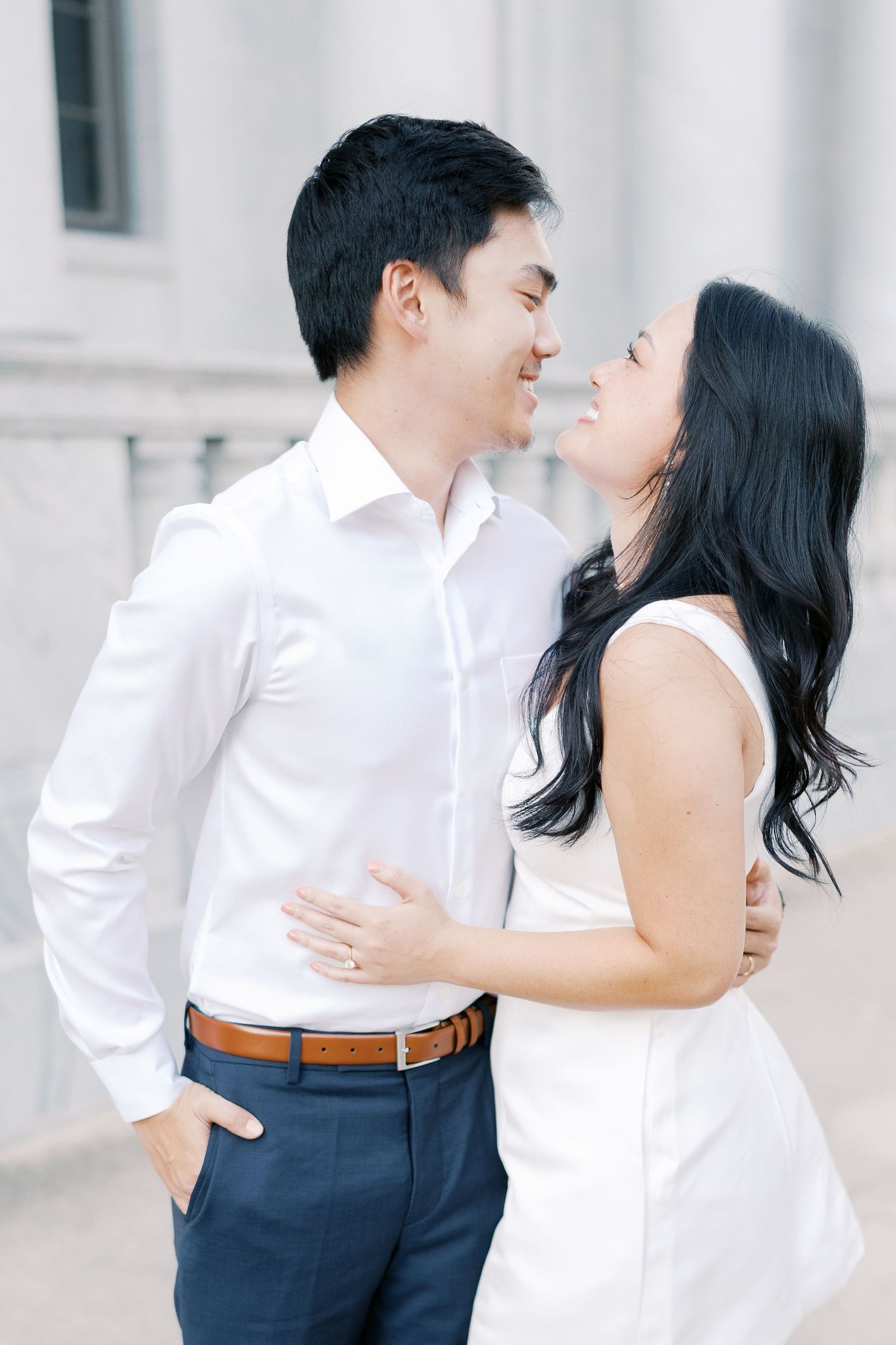 A romantic couple in white and navy blue outfits embrace in front of a light-colored building.