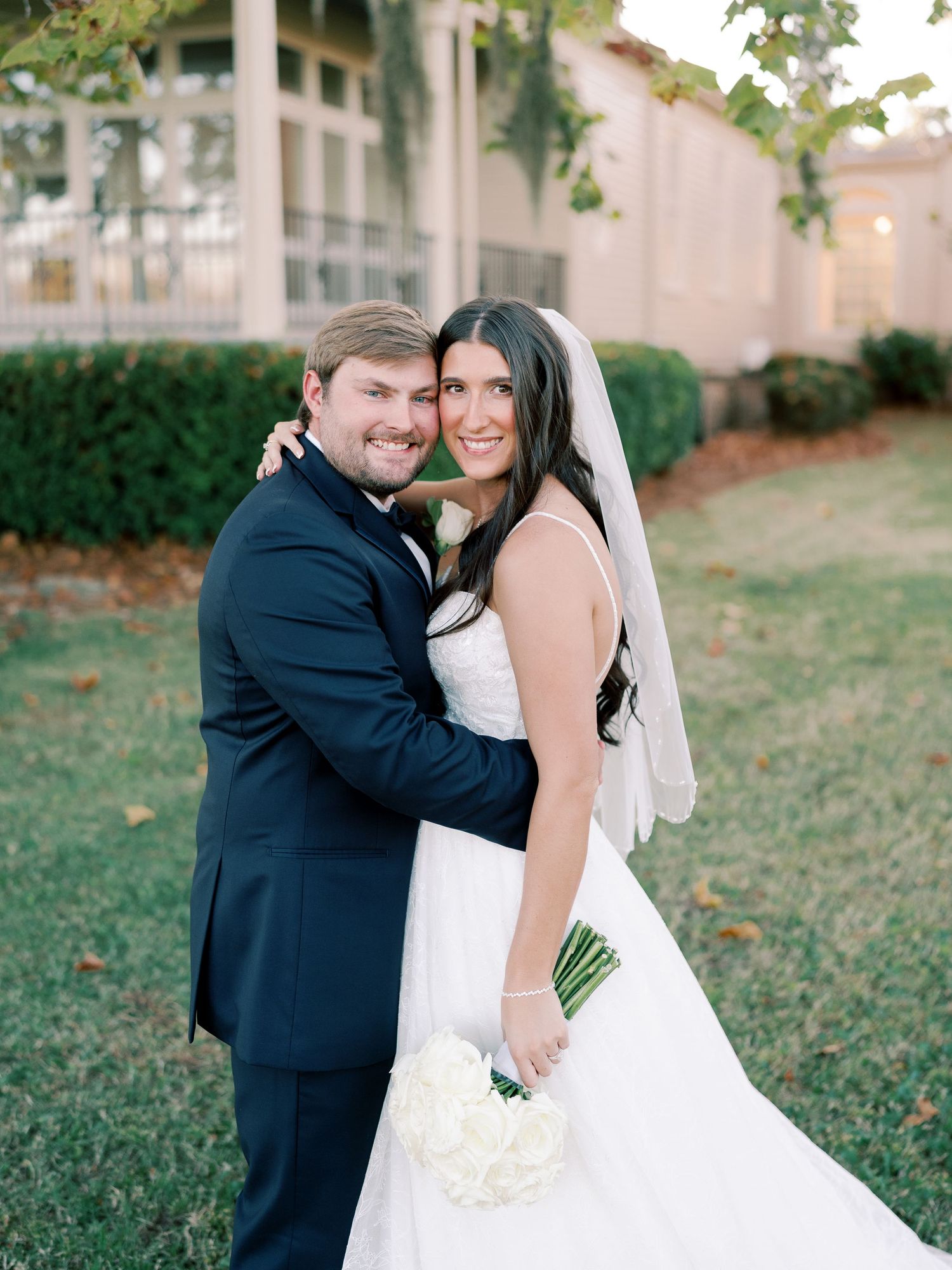 Happy wedding couple embraces on lawn in front of elegant building.