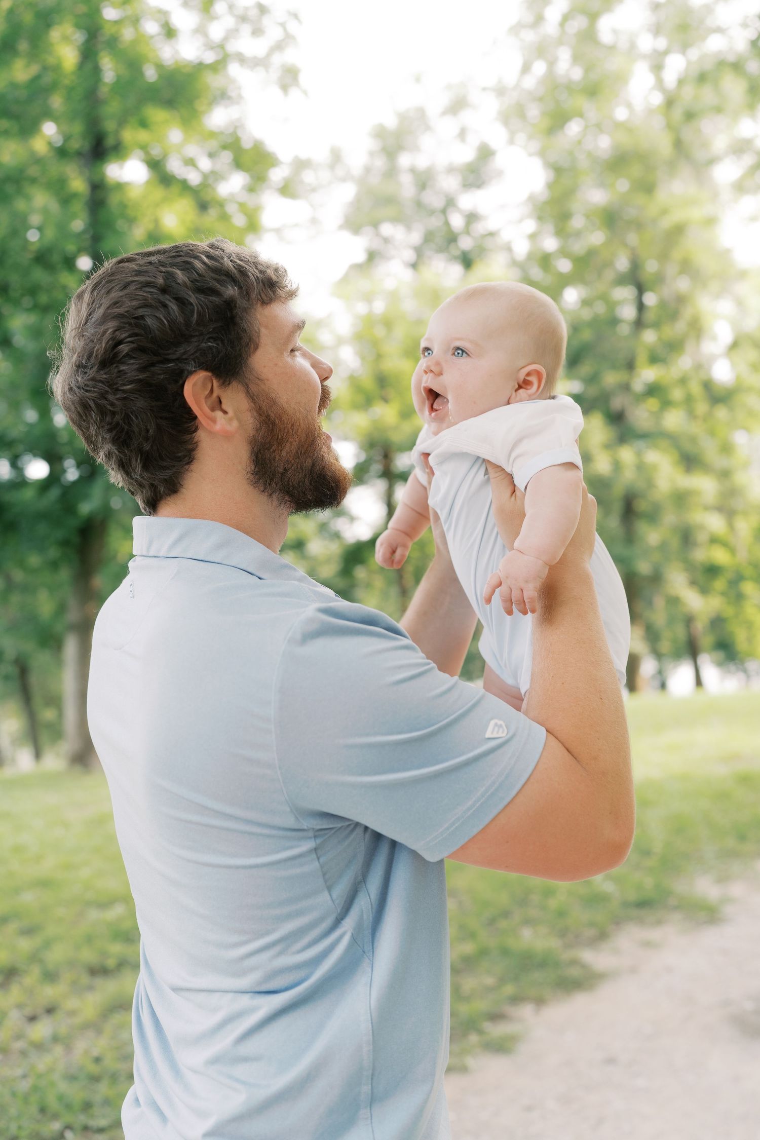 A parent in blue holds up a joyful baby against a green park background.