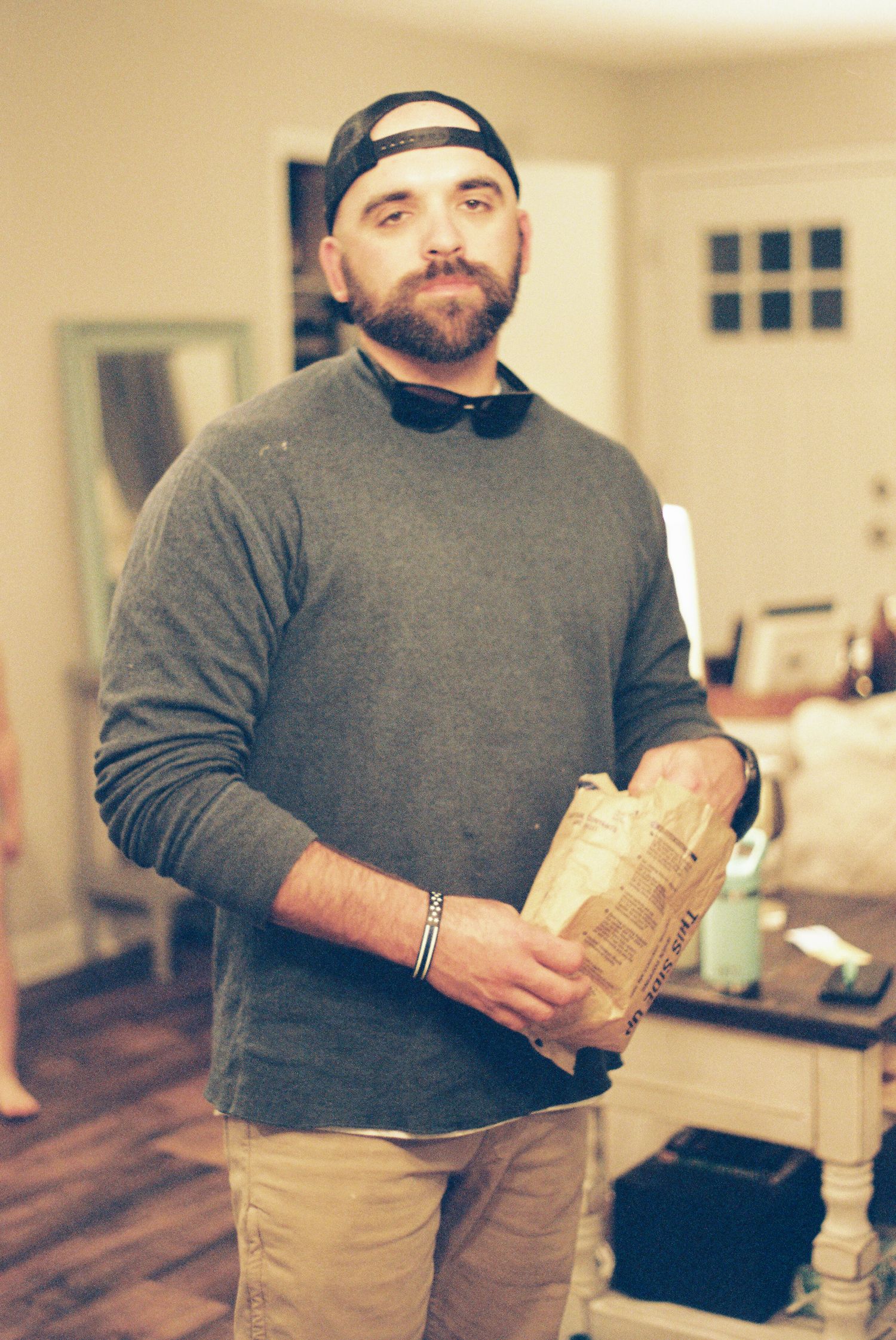 Person in a gray sweater and khaki pants holding a brown paper bag in an indoor setting.