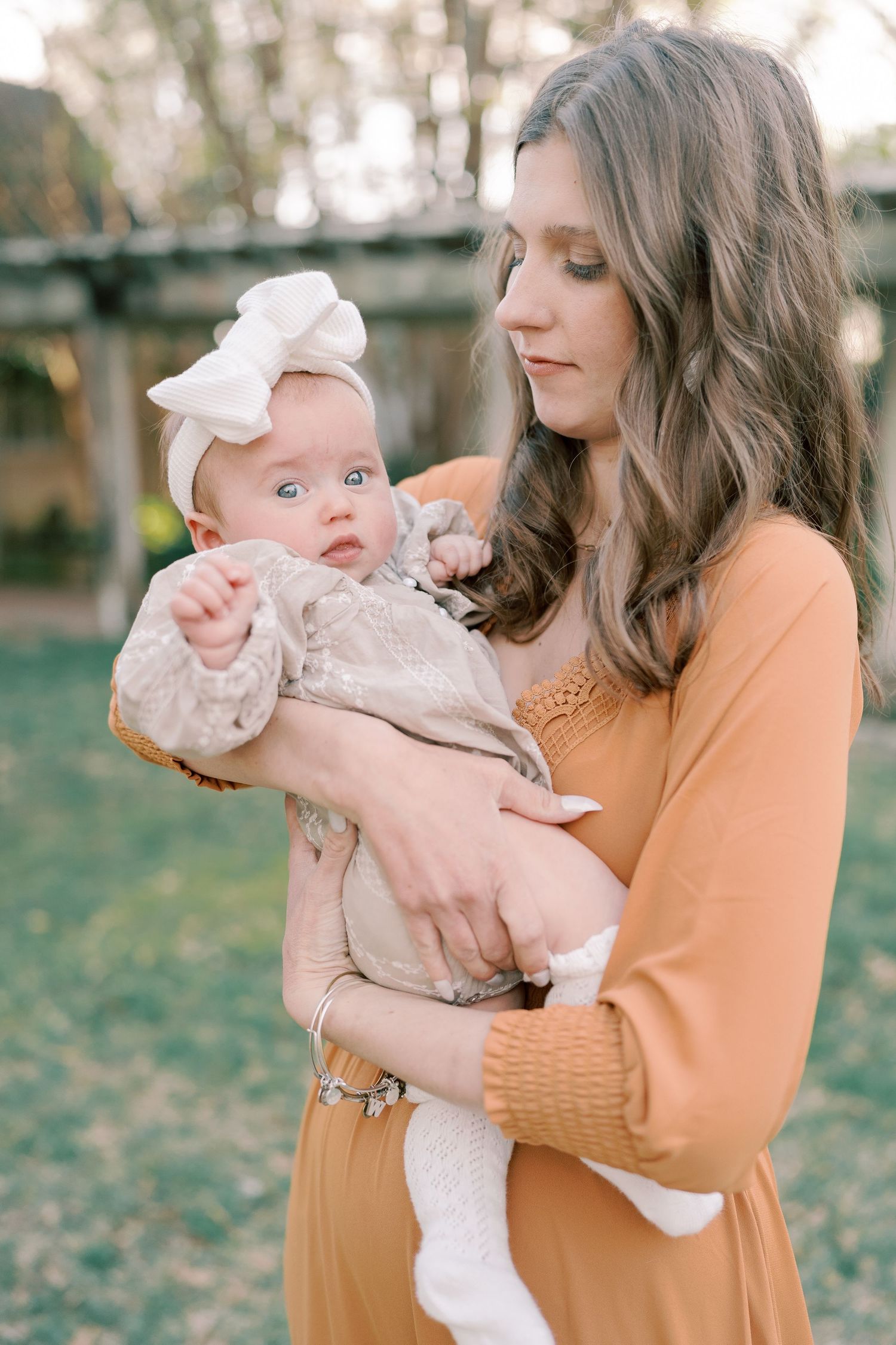 A baby wearing a white bow and dress is held in a warm autumn outdoor setting.
