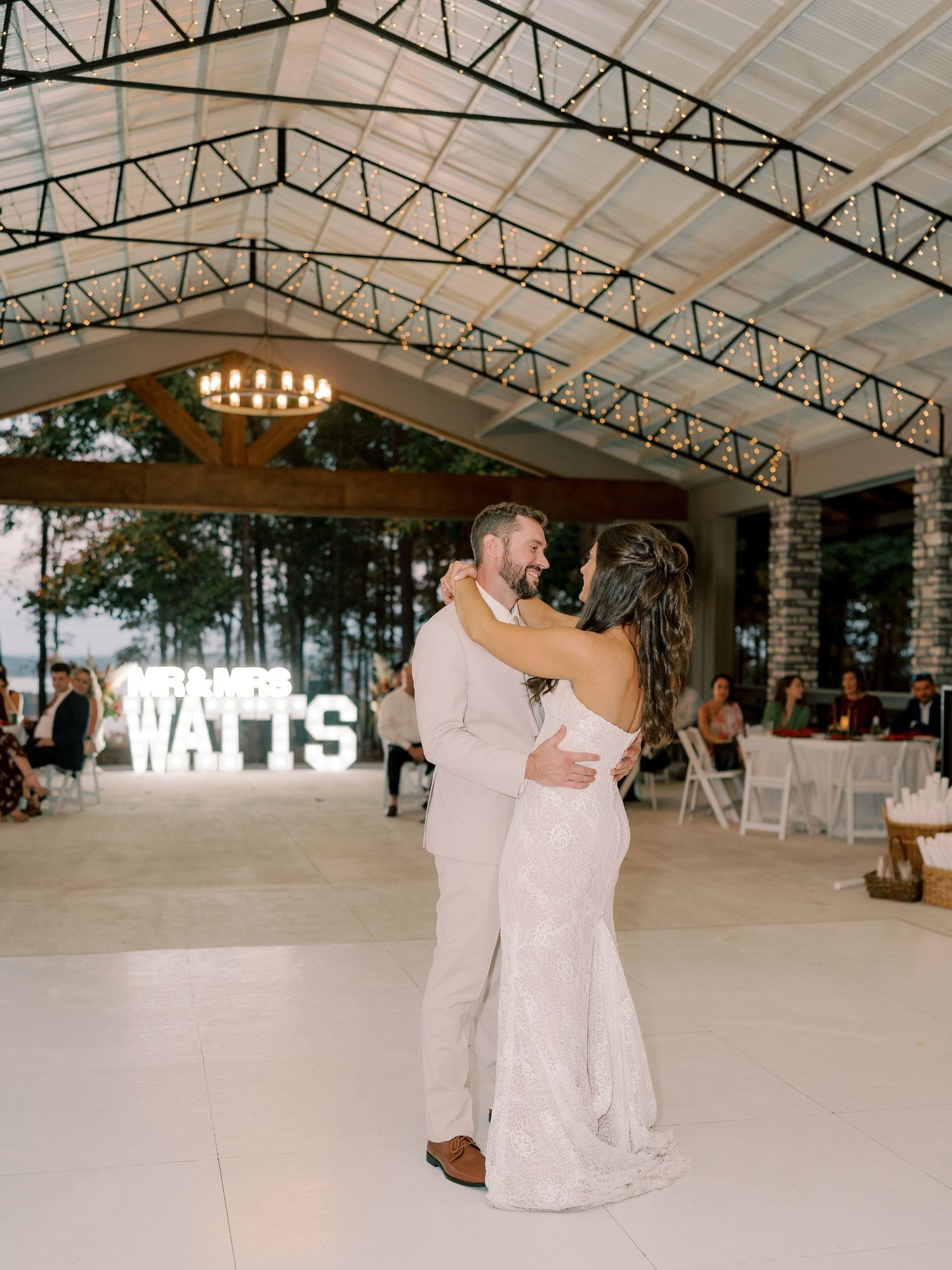 Newlyweds sharing first dance under covered reception venue with illuminated name sign.