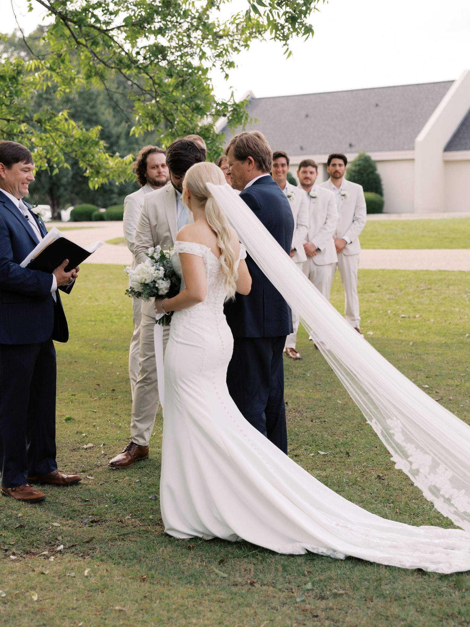 A wedding party gathers outdoors as a bride's long veil flows in the breeze.