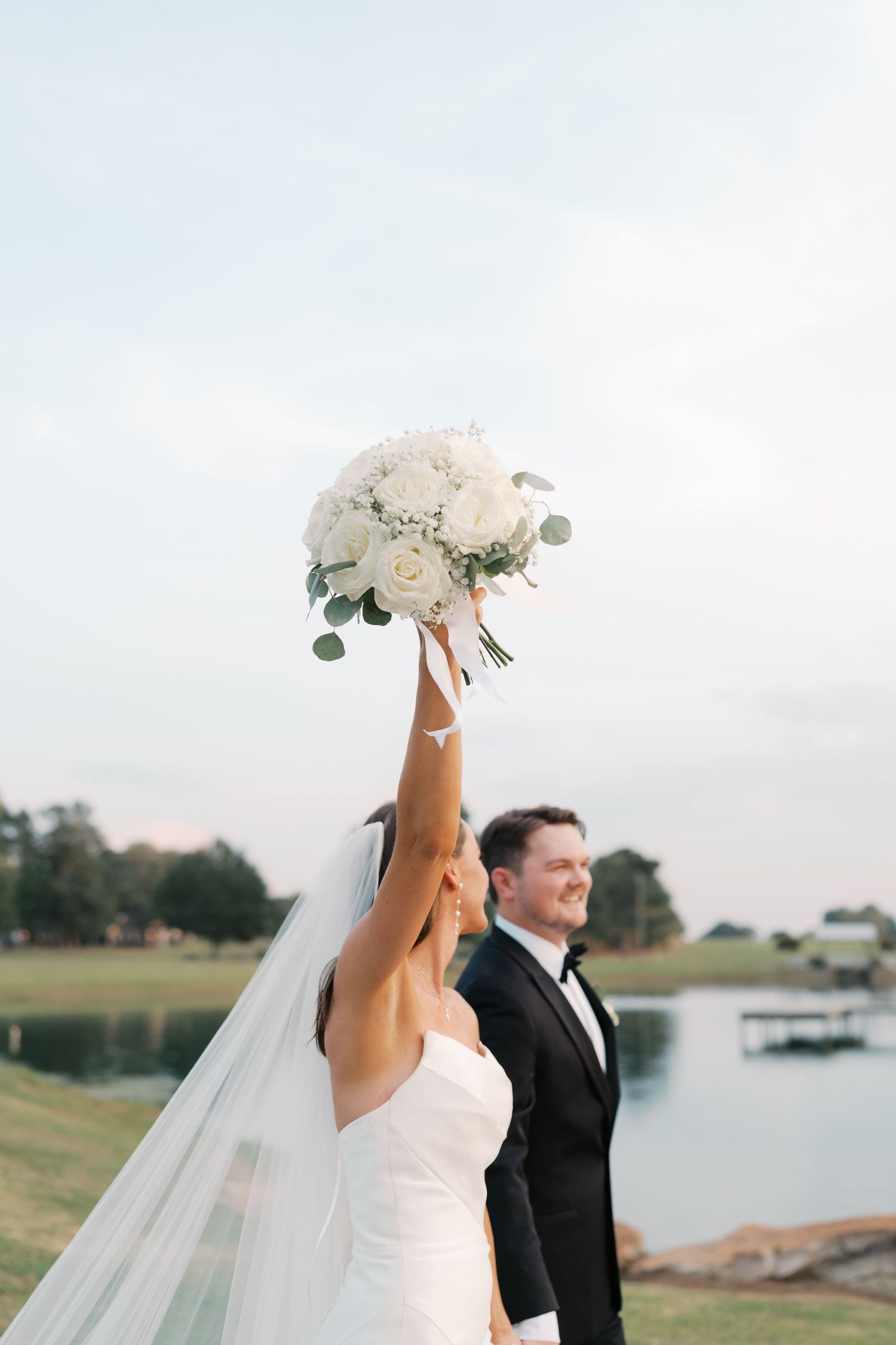 A romantic wedding couple poses for photos by a serene lakeside setting with white floral bouquet.
