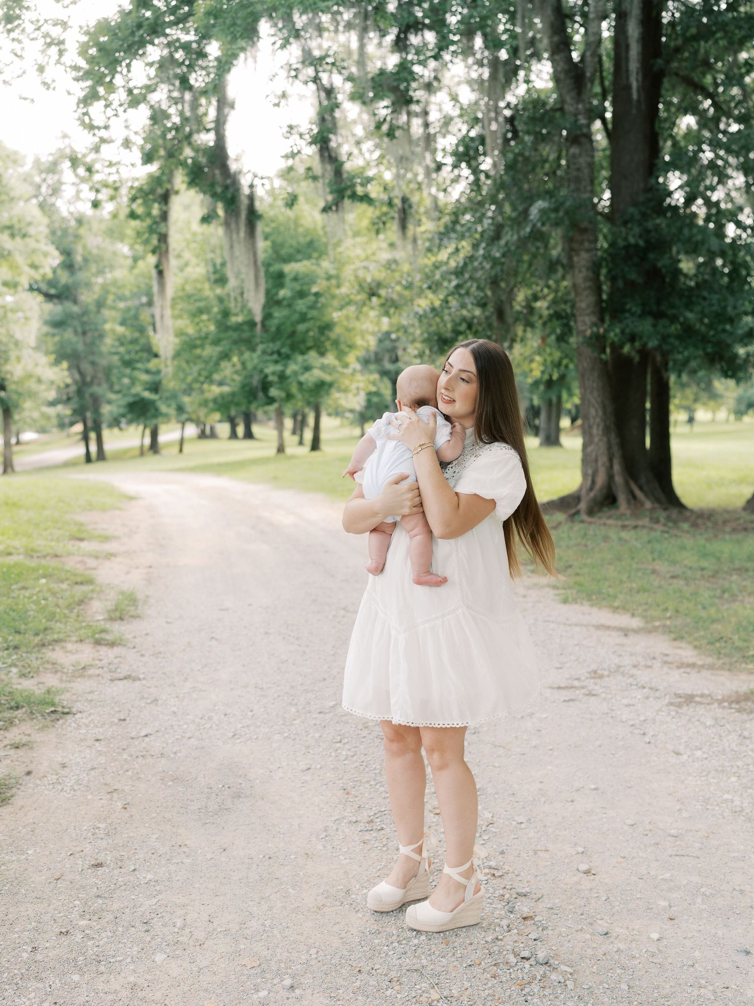 A woman in a white dress holds an infant while standing on a peaceful forest path.