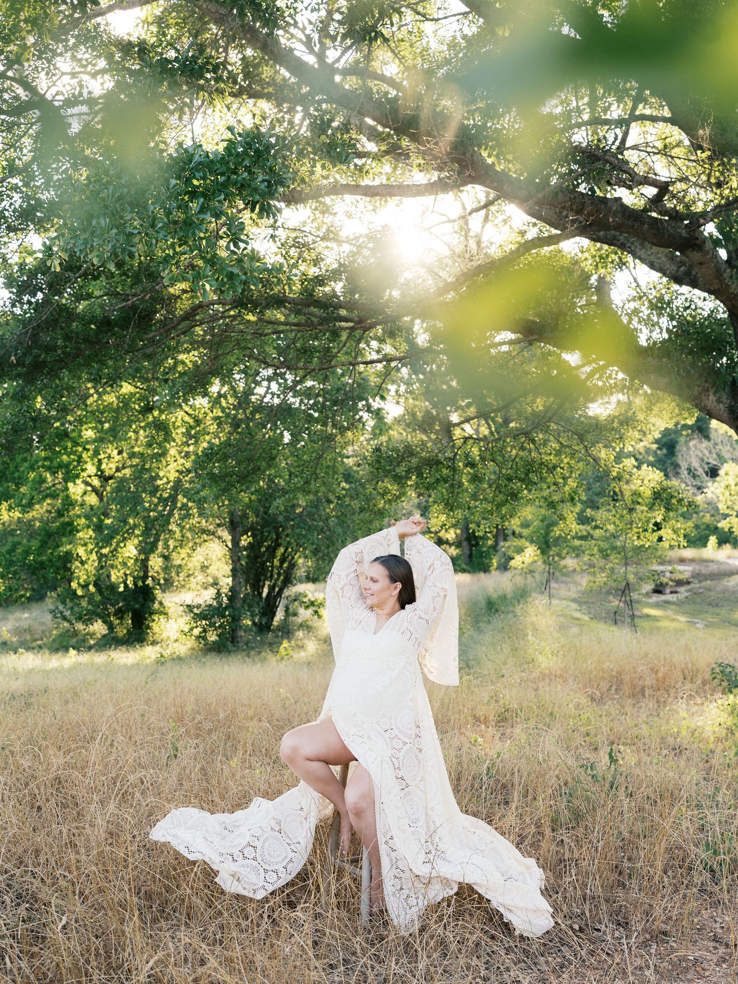 Artistic maternity portraits with billowing white lace dress in sunlit meadow during sunset.