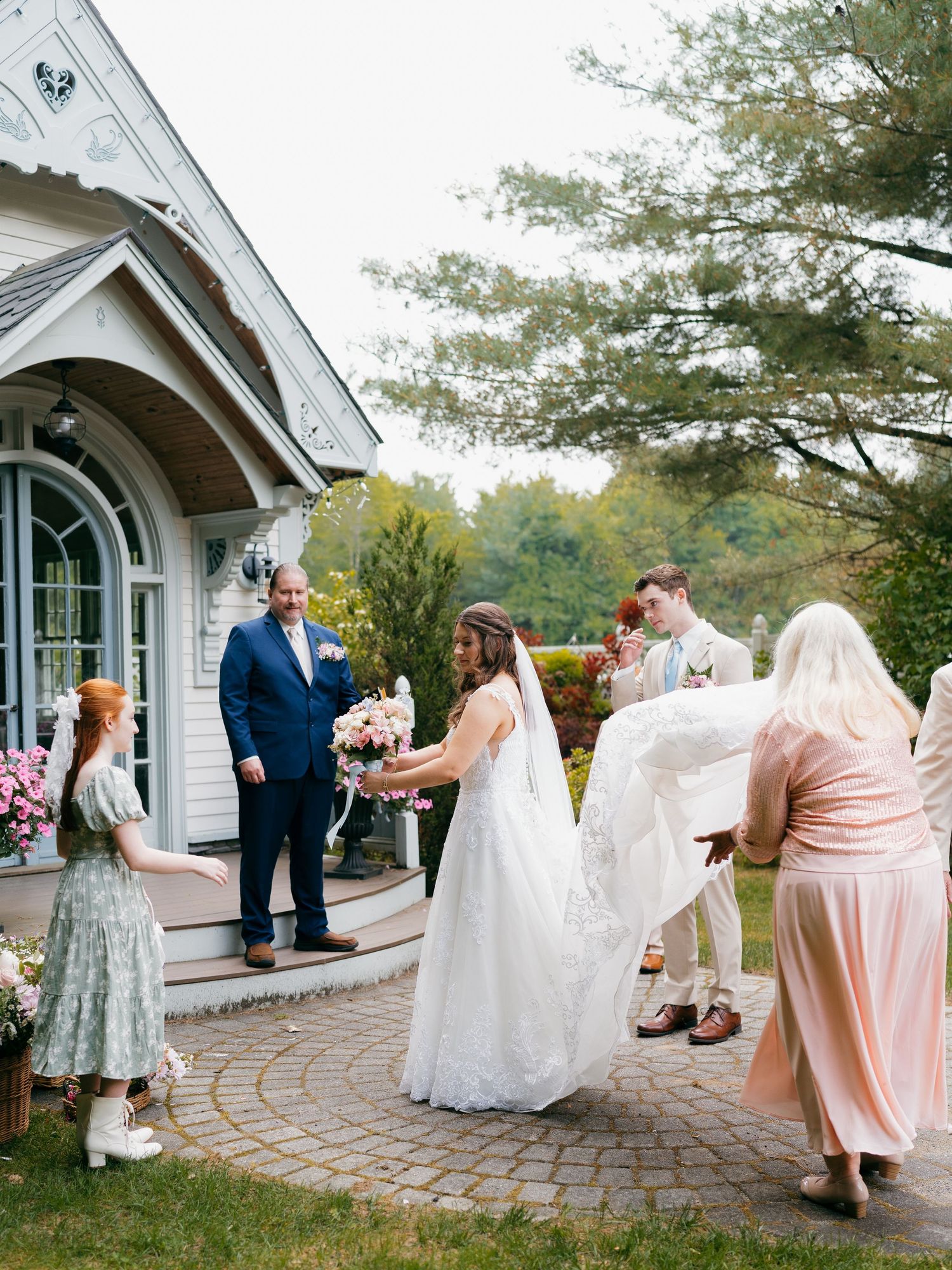 Bridesmaids in pastel dresses helping arrange the bride's flowing white gown on stone patio.