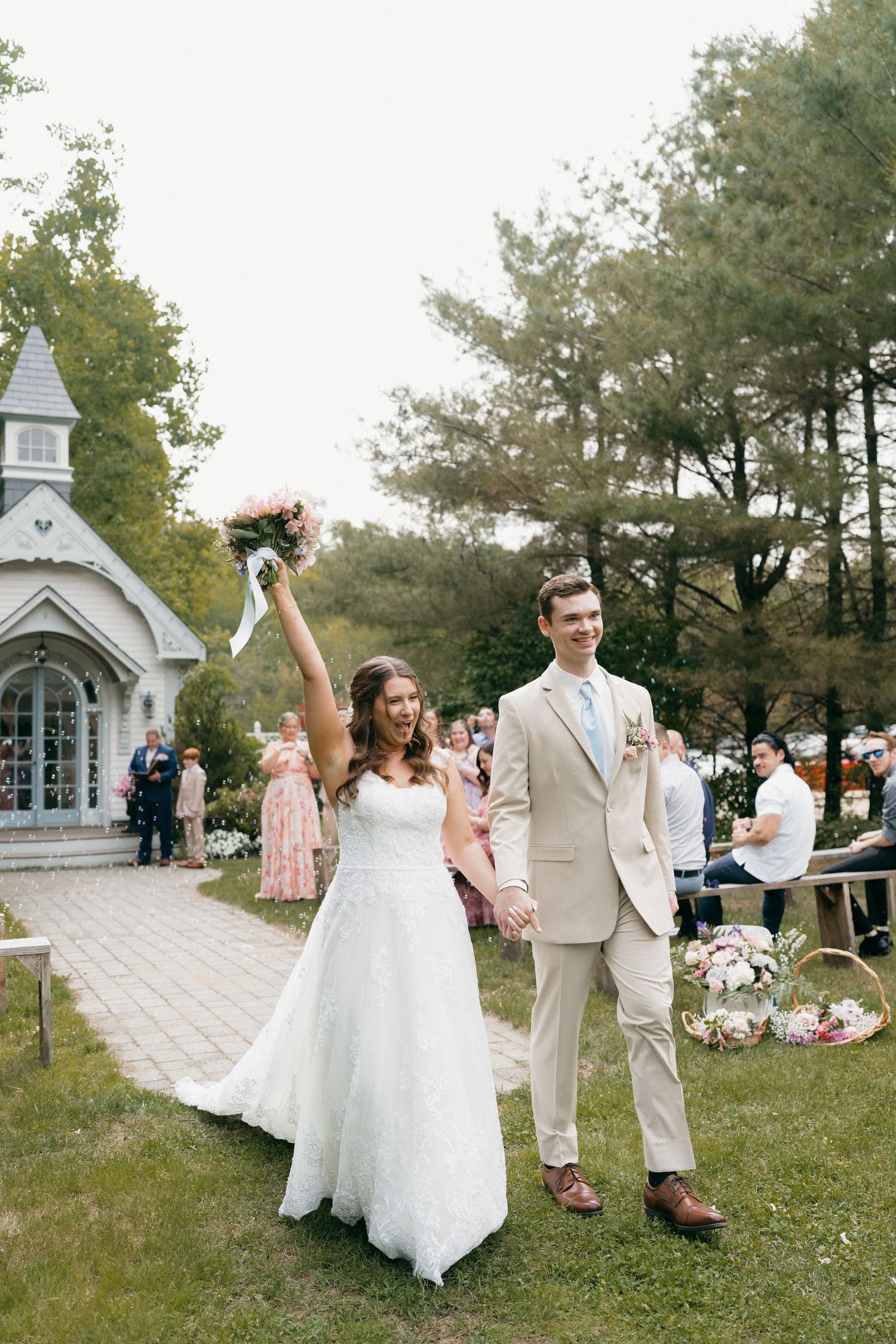Newlyweds celebrate outside a quaint white chapel with raised bouquet and joyful expressions.