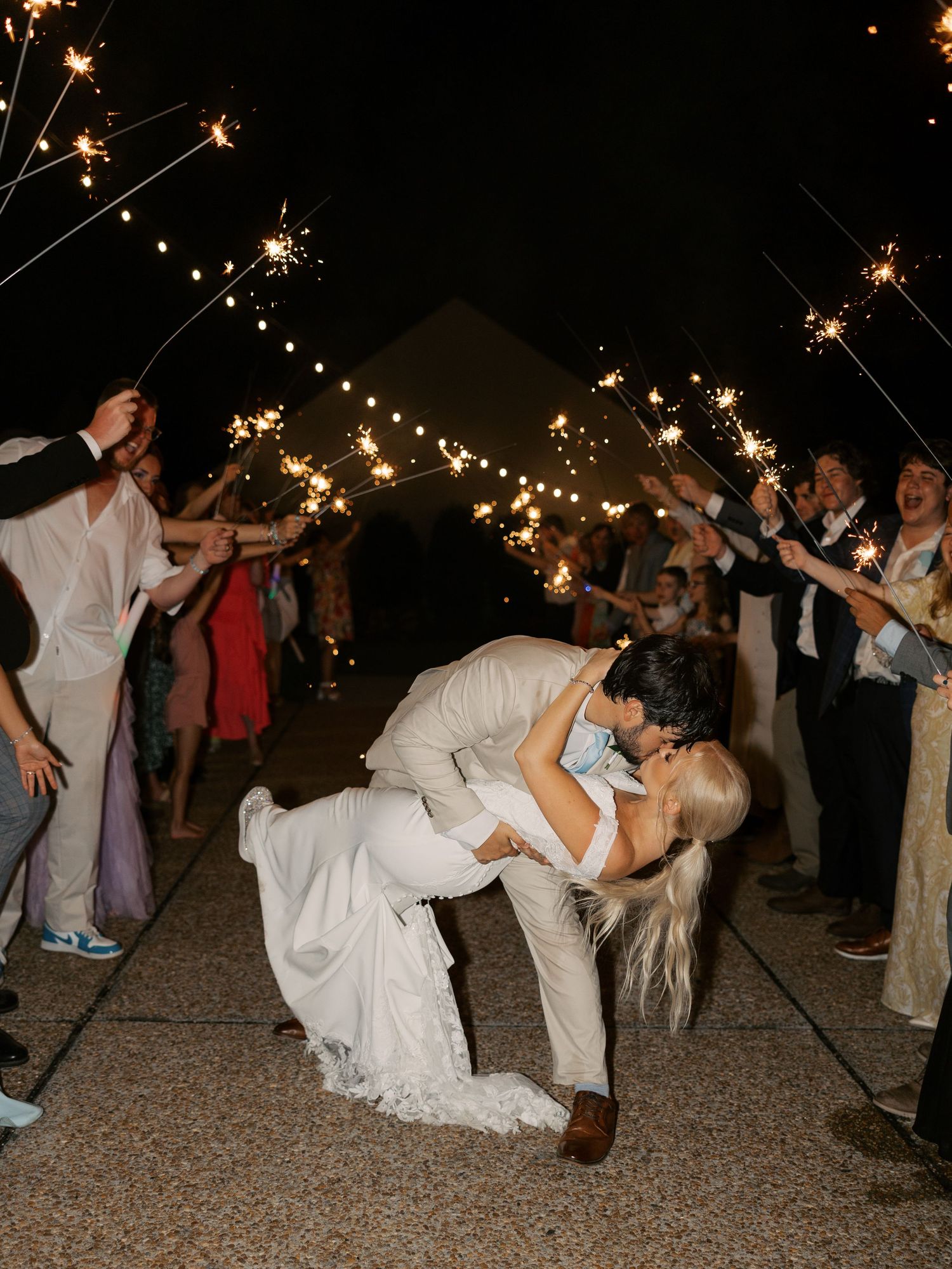 Dramatic dip kiss moment during wedding sparkler exit surrounded by celebrating guests.