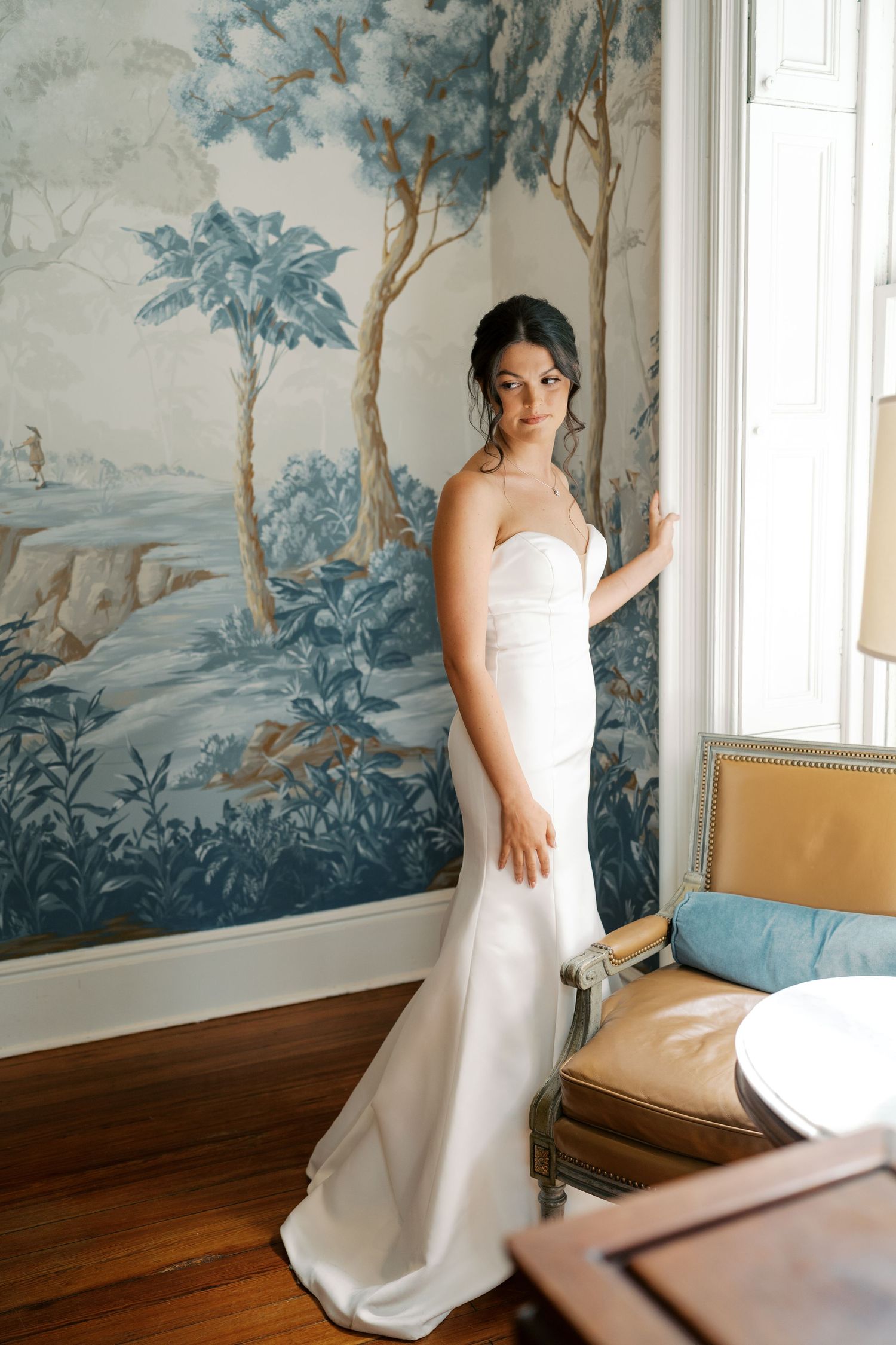 Bride in white dress poses against blue floral wallpaper in an elegant room.