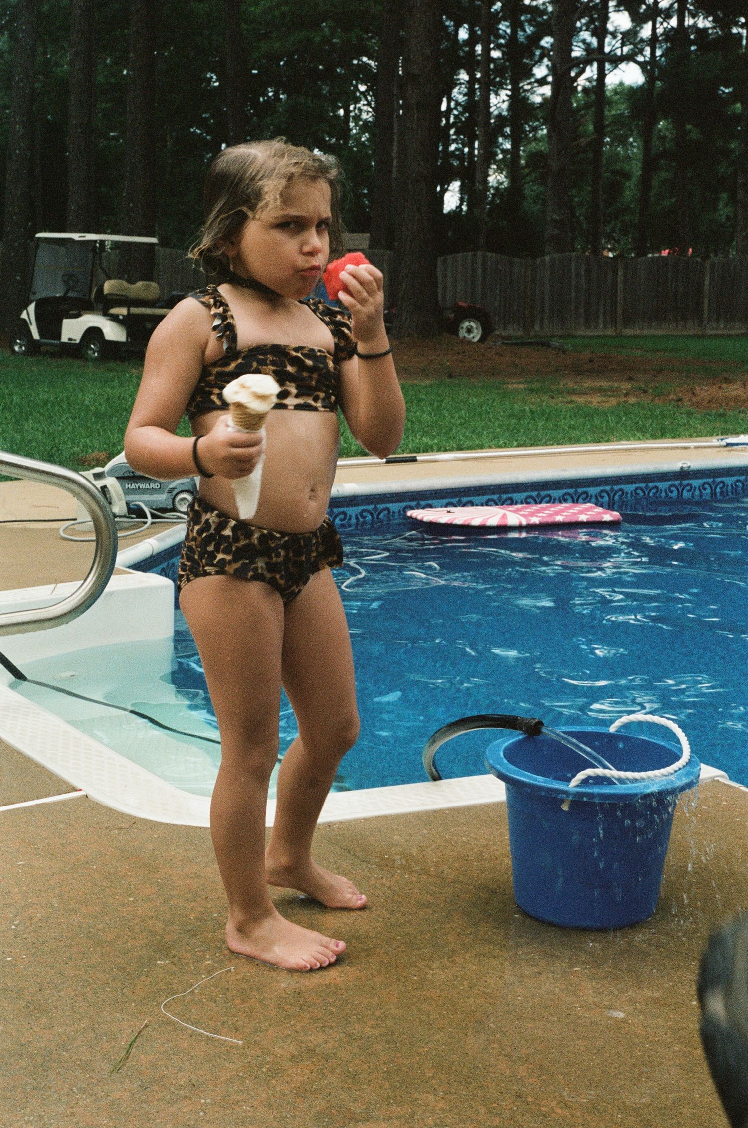 Small child playing near a swimming pool with blue buckets on concrete deck.