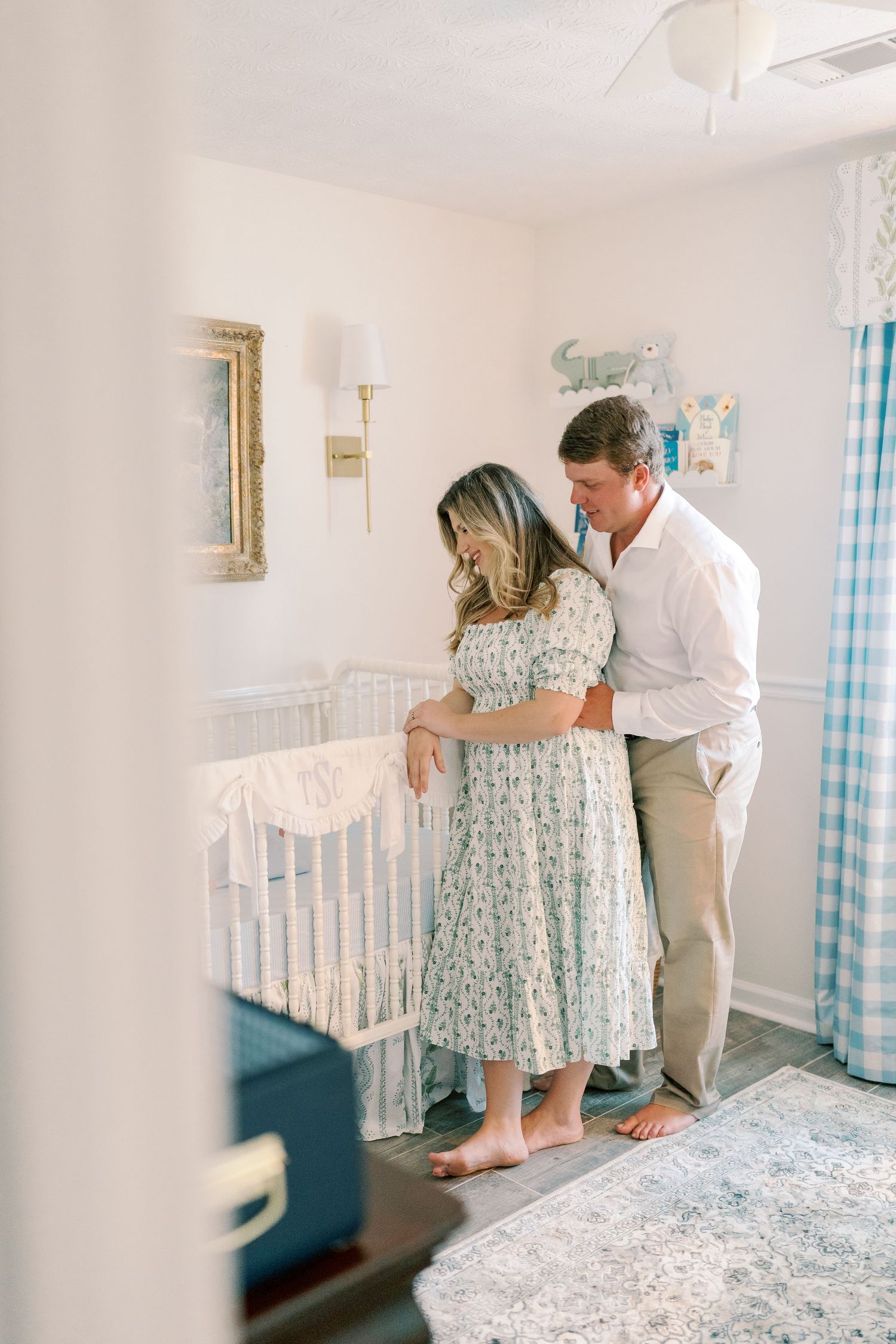 Expecting couple embraces near a white crib in a nursery with blue checkered curtains.