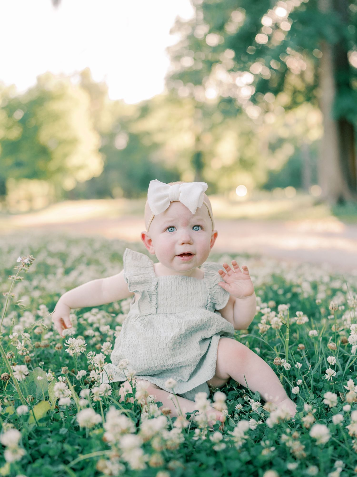 Baby in grey romper sitting among blooming white wildflowers in a sunlit field.