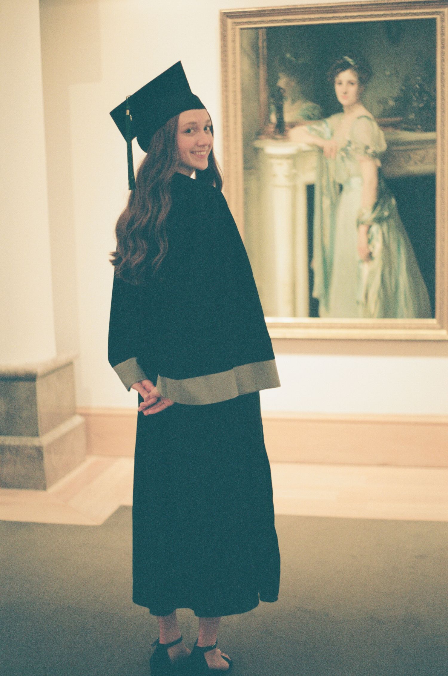 Graduate in black cap and gown walking through an art gallery with framed paintings.