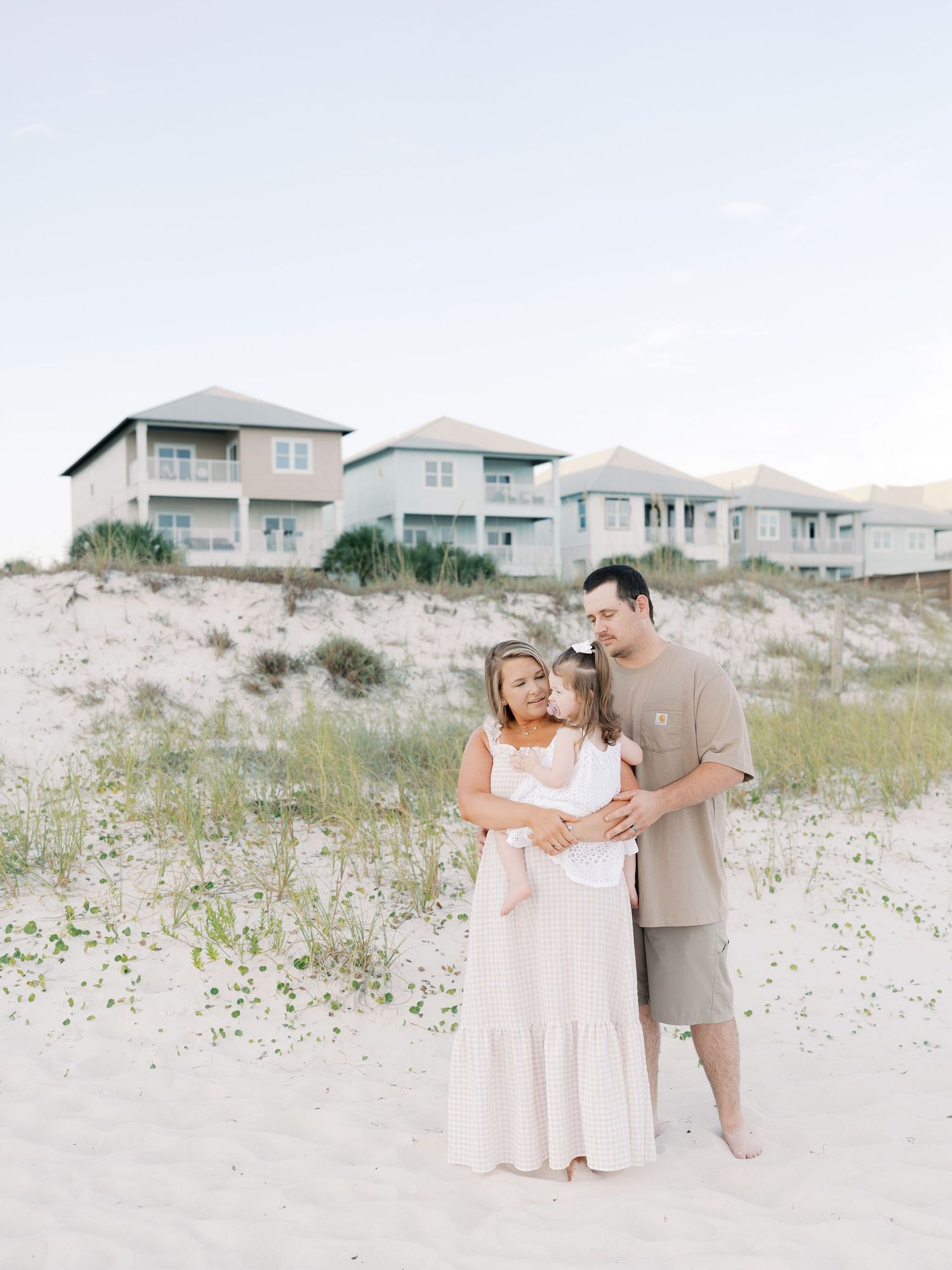 Family enjoying a beach photoshoot with coastal homes visible on sand dunes in the background.