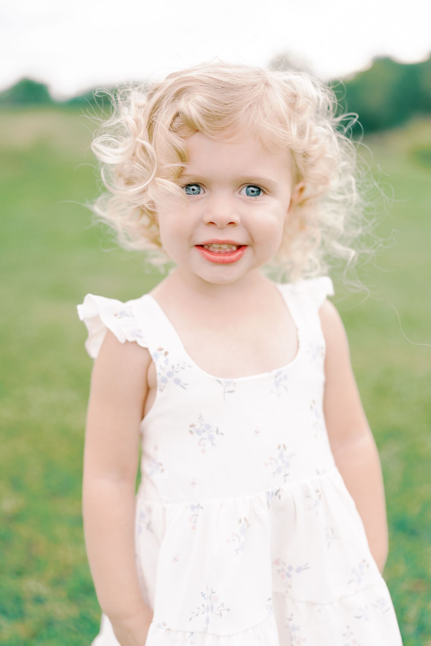 Sweet toddler in white lace dress playing outdoors in bright green field.