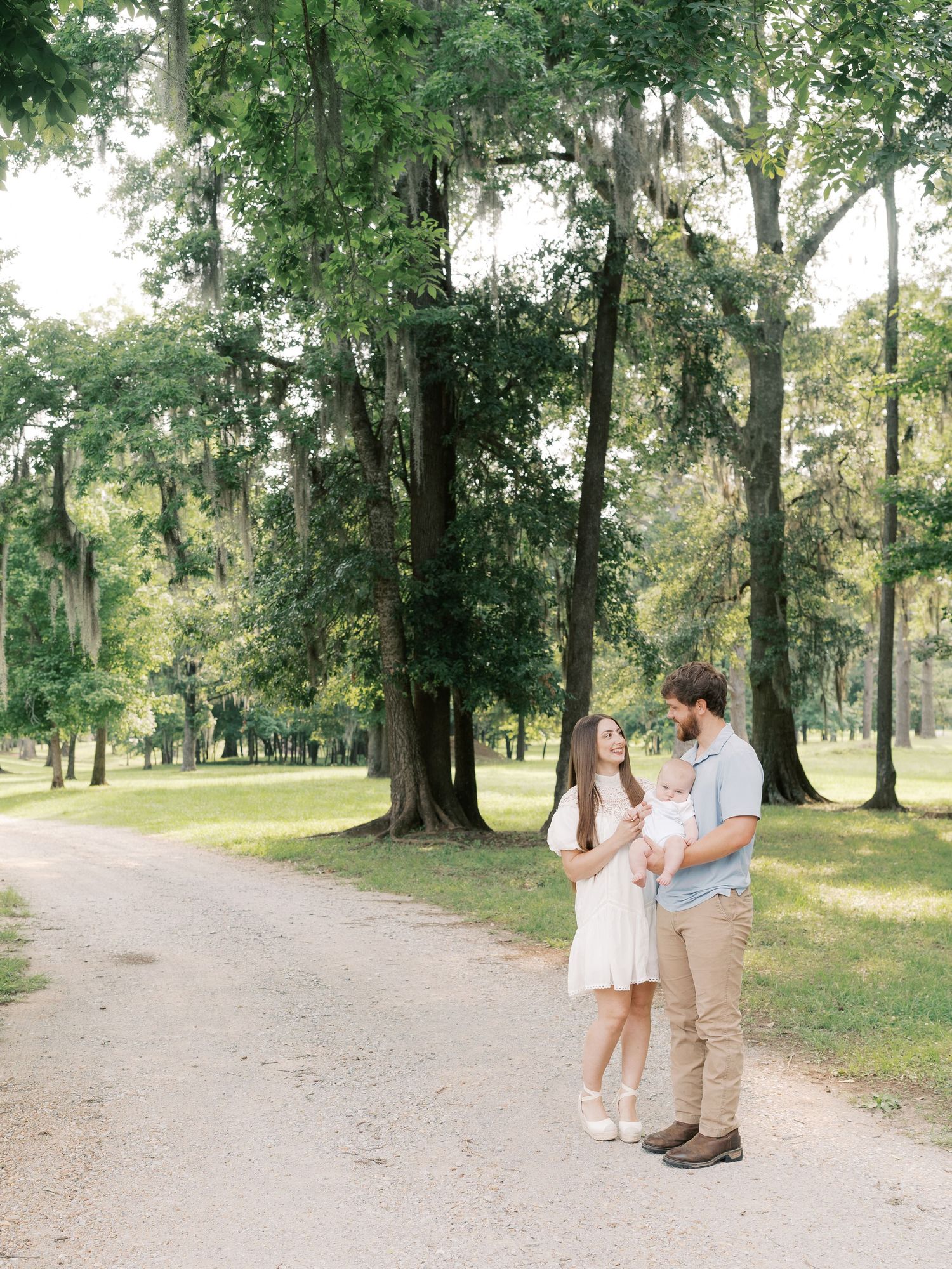 A couple and baby stroll down a peaceful wooded trail in matching light-colored outfits.