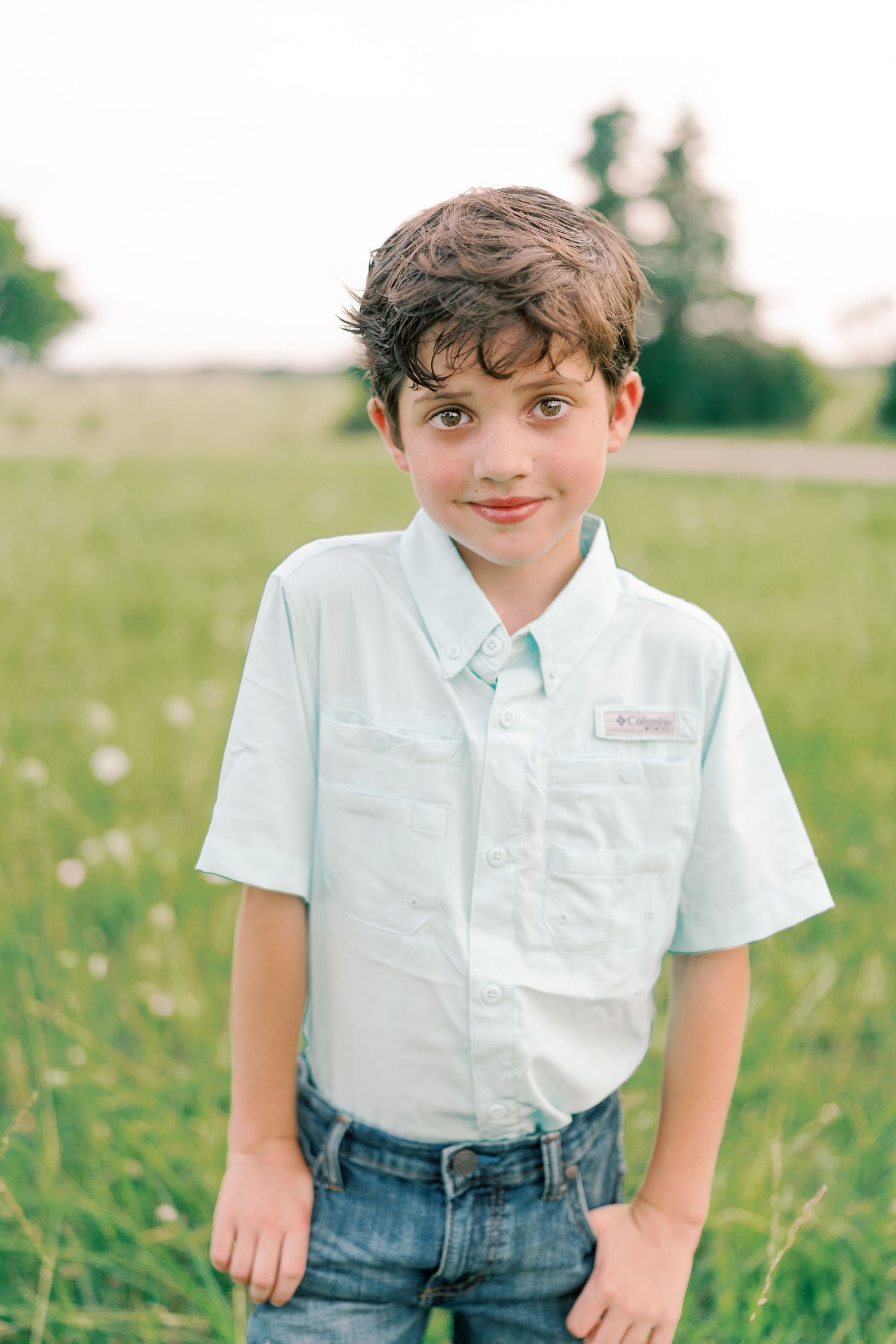 Child in white polo shirt and jeans poses in a green summer field.