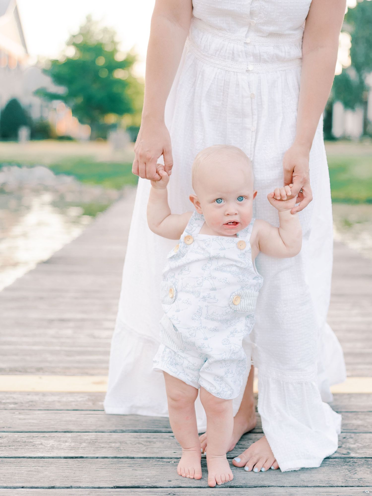 Baby taking first steps on wooden dock while holding parent's hands.