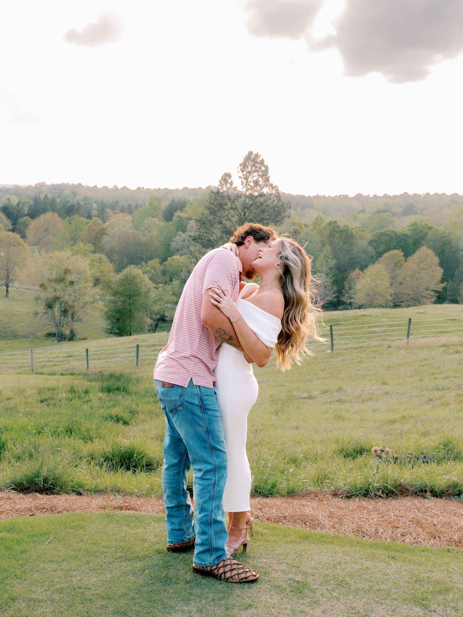 Romantic dip pose on grassy hilltop with scenic countryside backdrop.