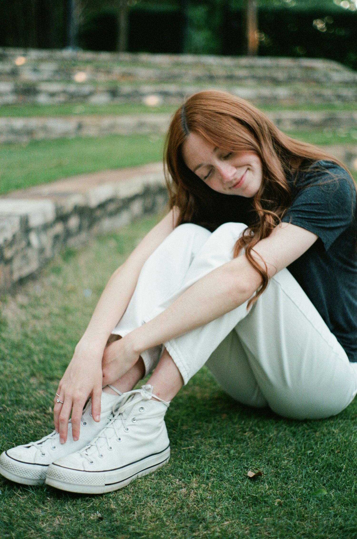 Person in black top and white pants relaxing on grass while wearing white Converse sneakers.