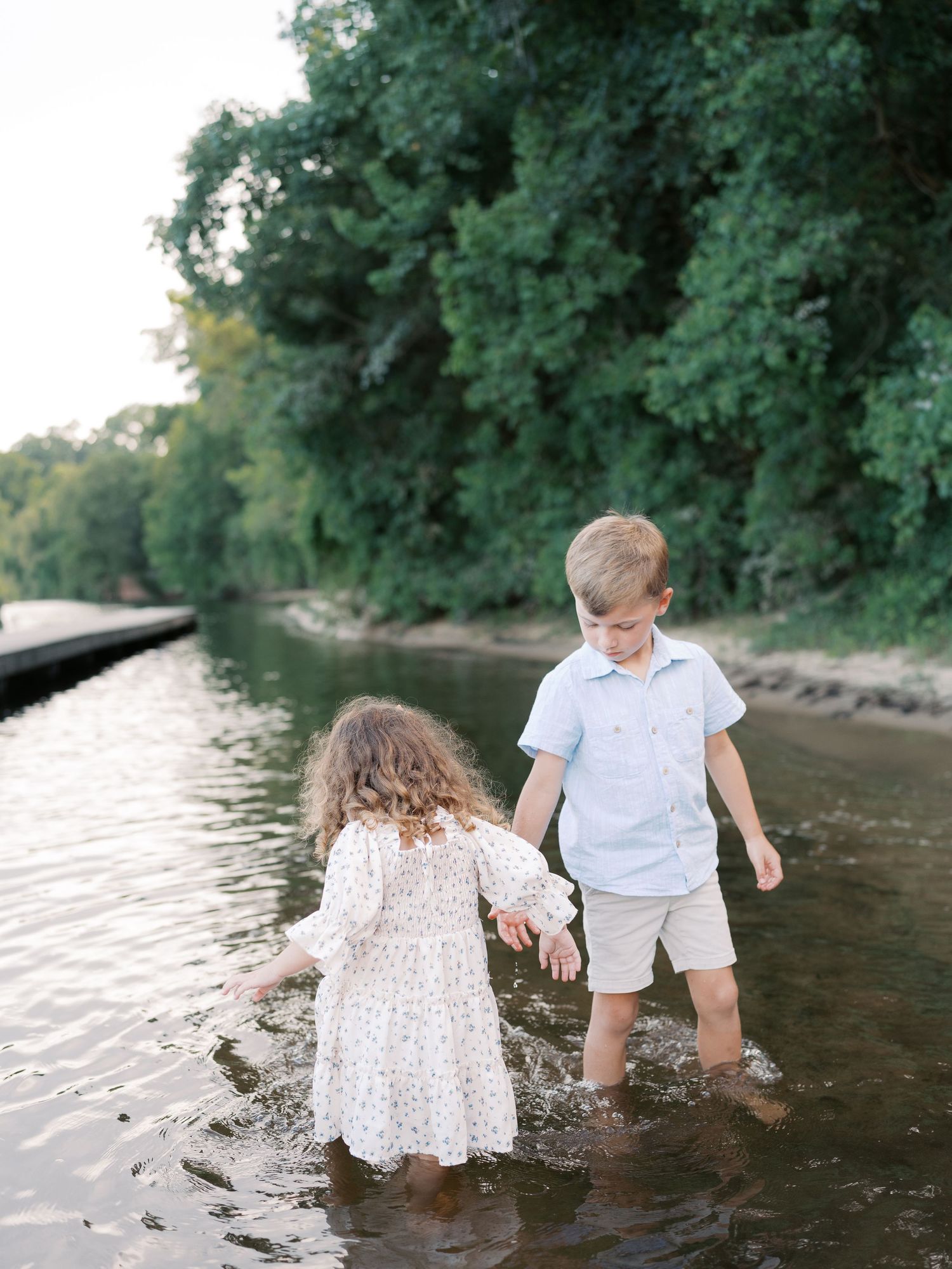 Children playing and splashing in shallow water at the edge of a wooded riverbank.