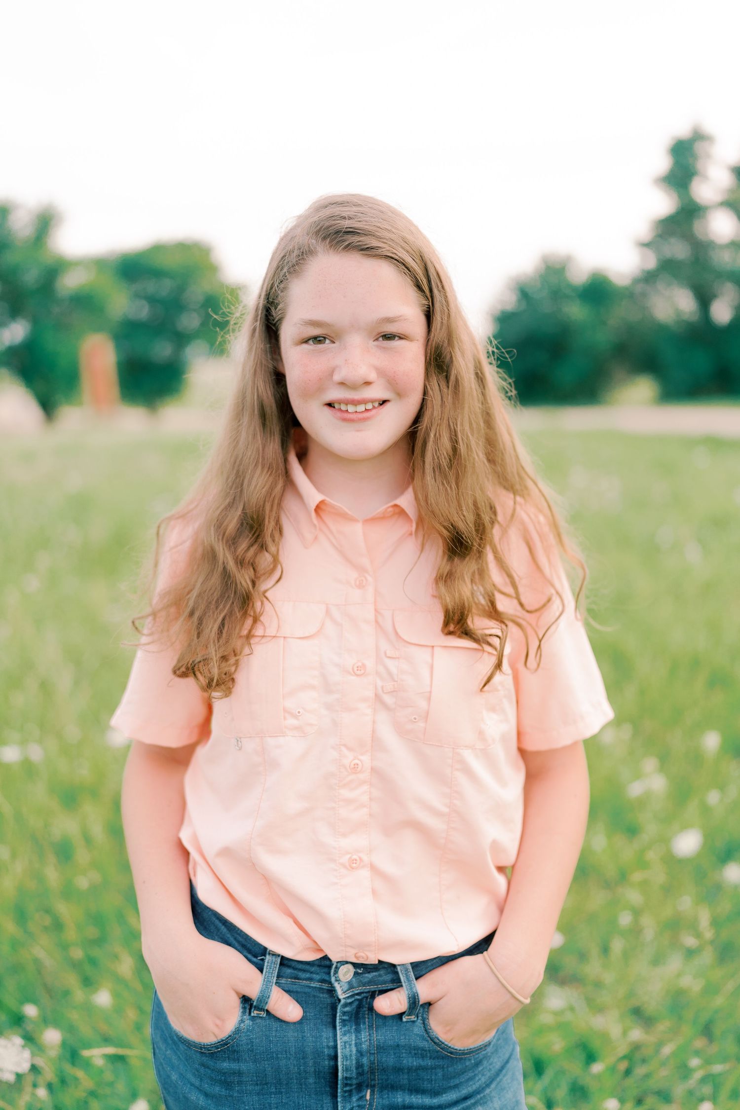 Person wearing peach colored shirt and jeans poses in a summer meadow.