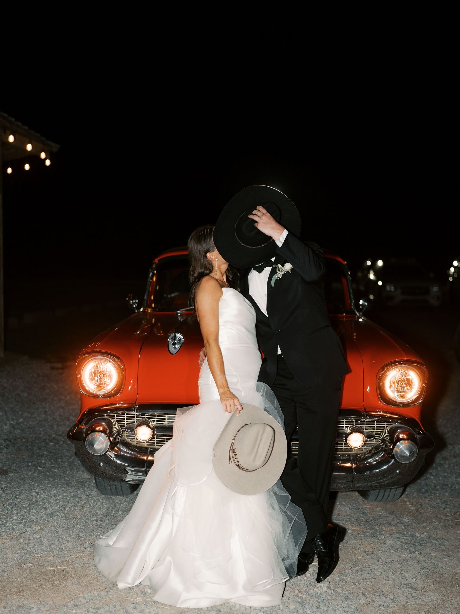 A bride and groom pose romantically against a vintage red car at night.
