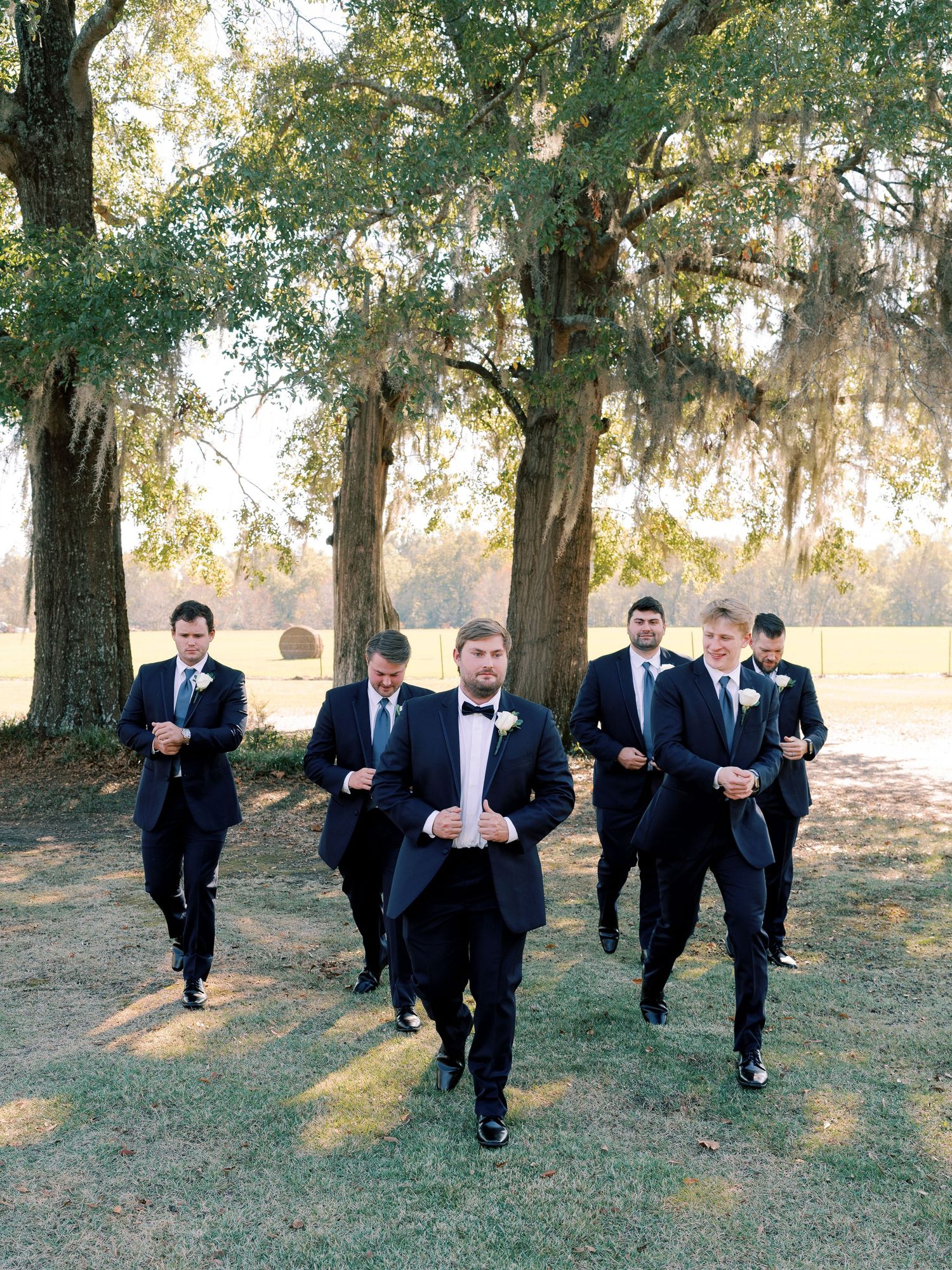 Wedding party in formal attire runs playfully across a grassy field on a sunny day.