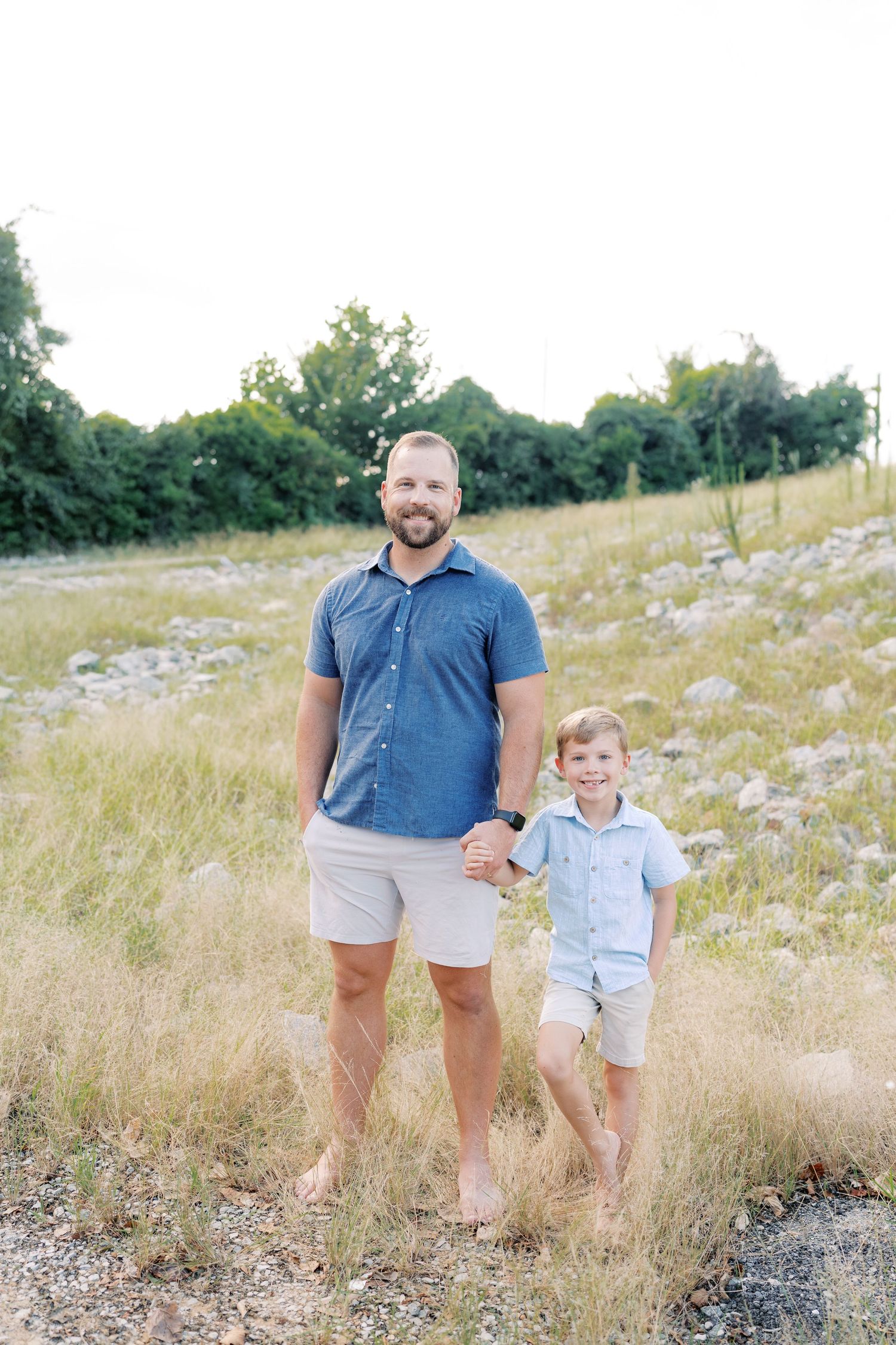 Father and son standing together barefoot in a grassy field on a sunny day.