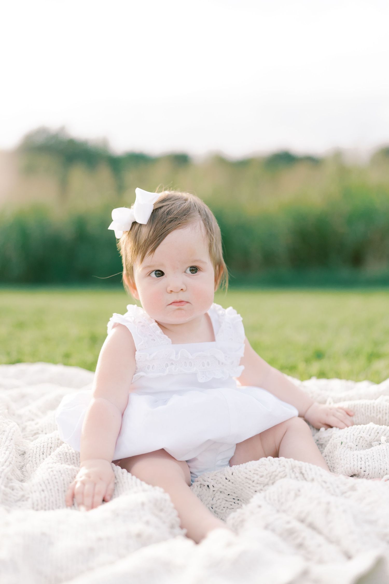 Baby in white dress poses on white blanket during outdoor portrait session.