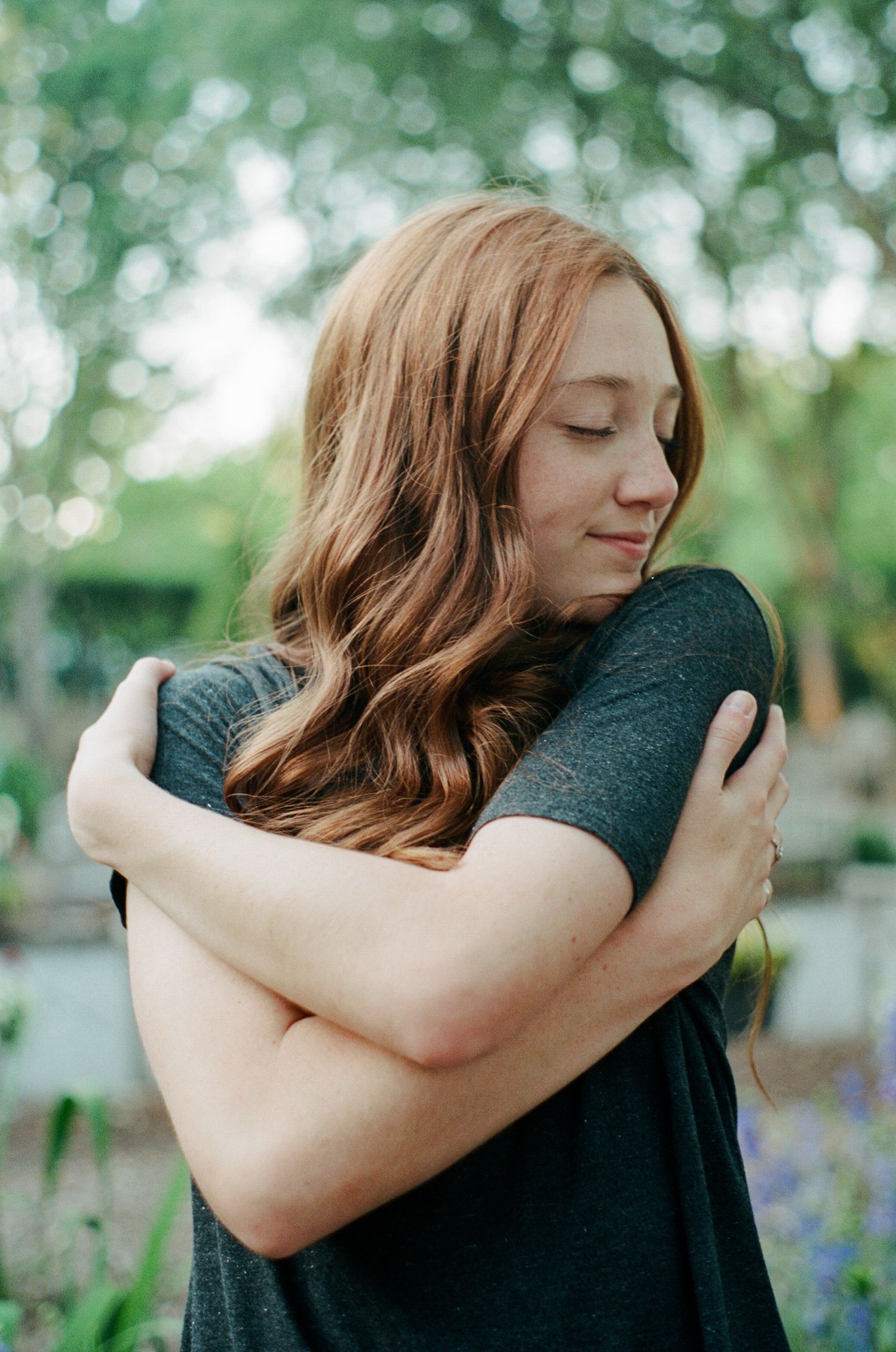 Person in dark clothing embracing themselves in a peaceful garden setting with bokeh background.