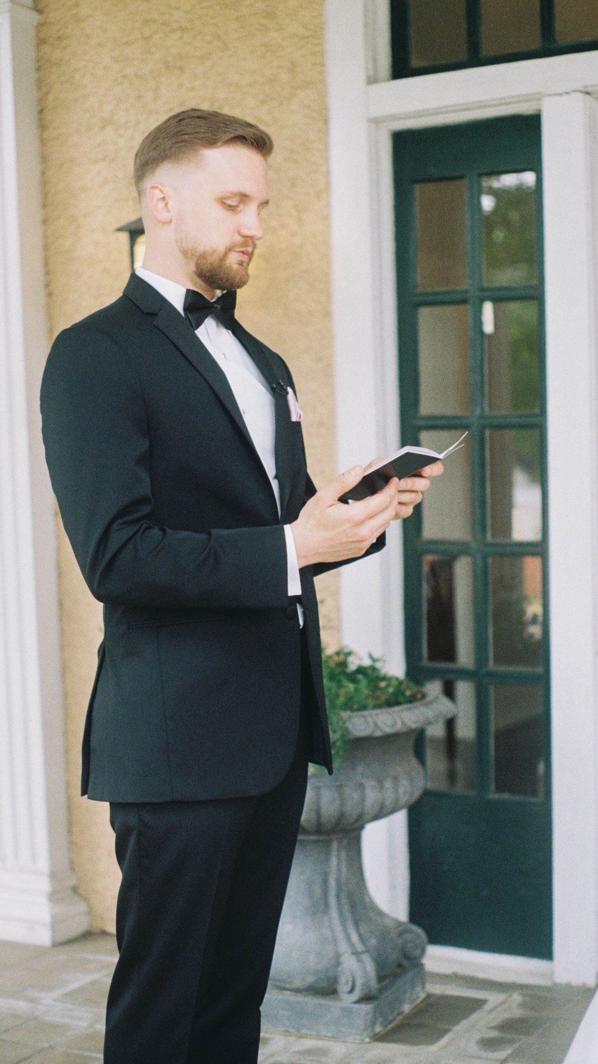 Person in formal attire checking phone outside elegant building entrance.