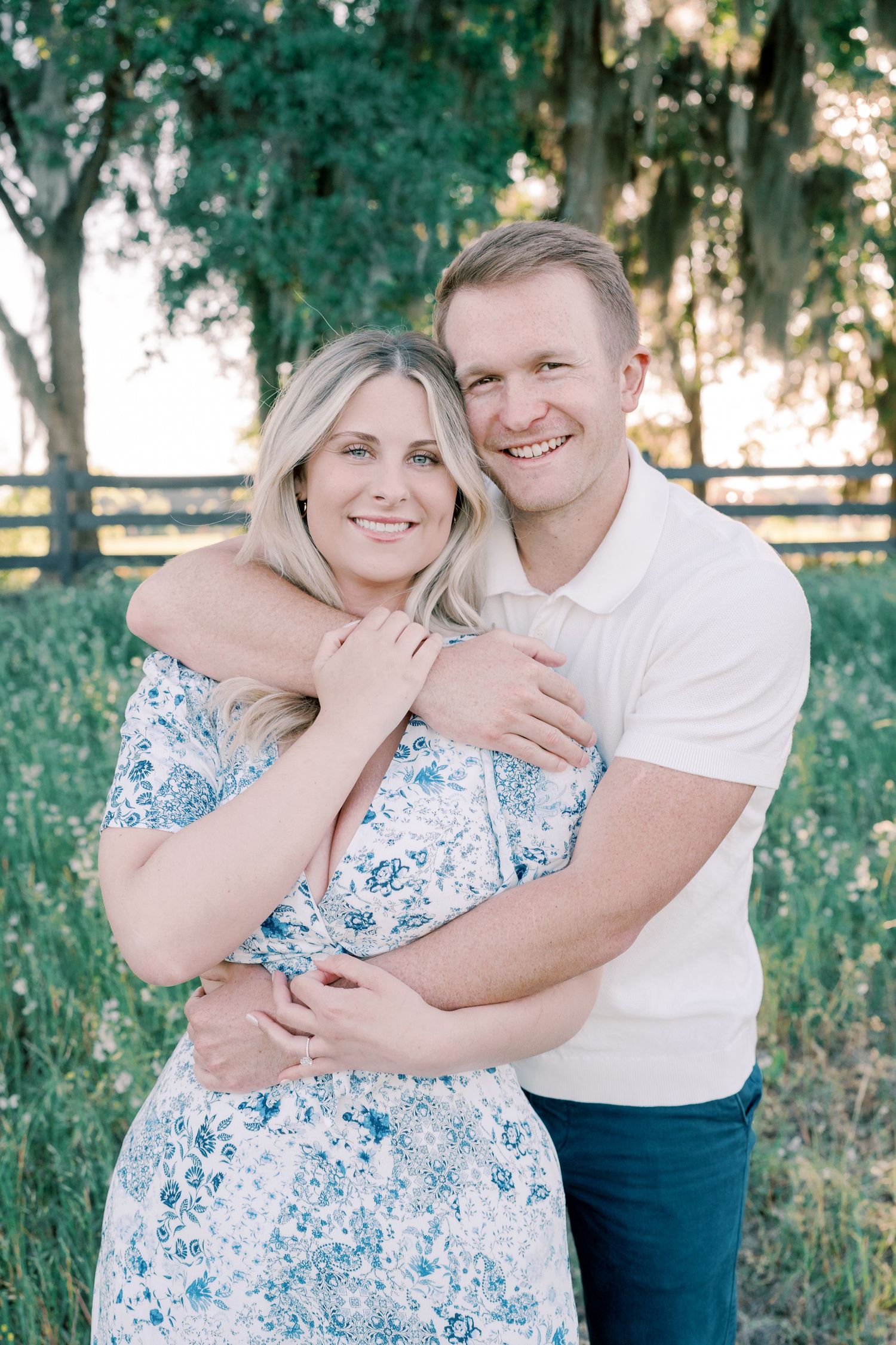 A couple embraces in a romantic outdoor photoshoot with trees and a wooden fence in the background.