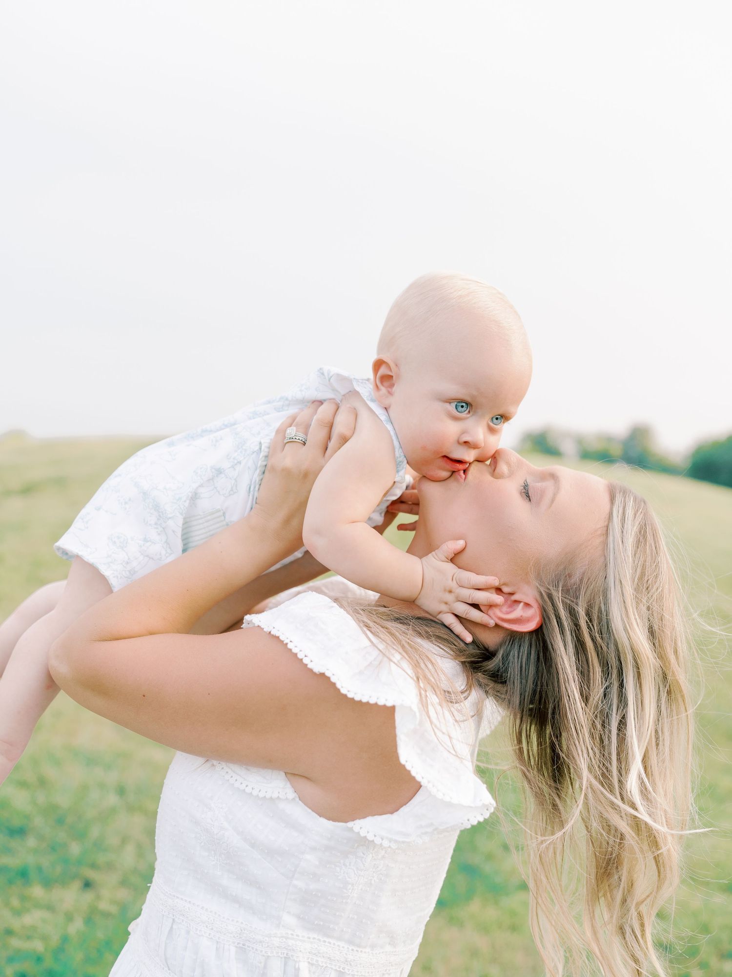 A tender moment of affection and play between parent and baby in white outfits against a sunny outdoor backdrop.