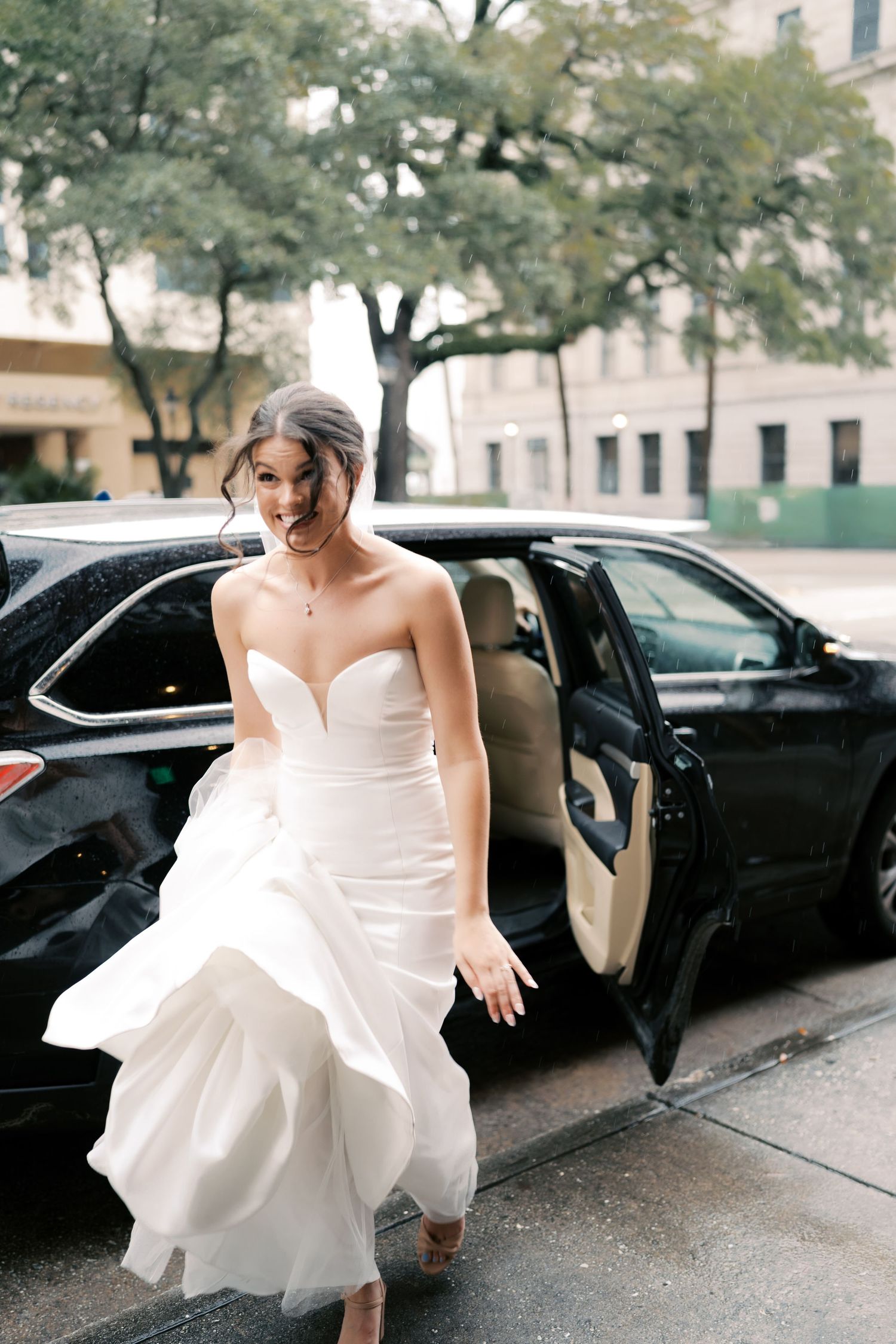 A bride in a white gown steps out of a black car on a city street.