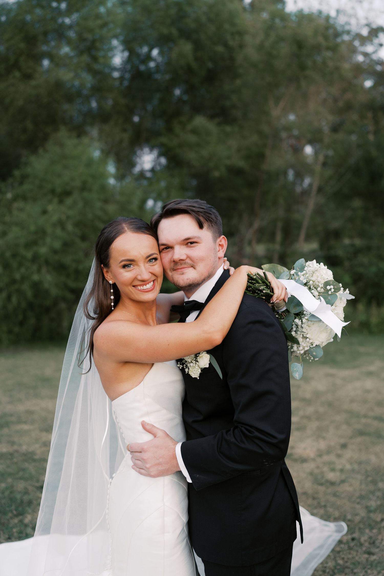 Wedding couple embracing outdoors in formal attire with white bouquet.