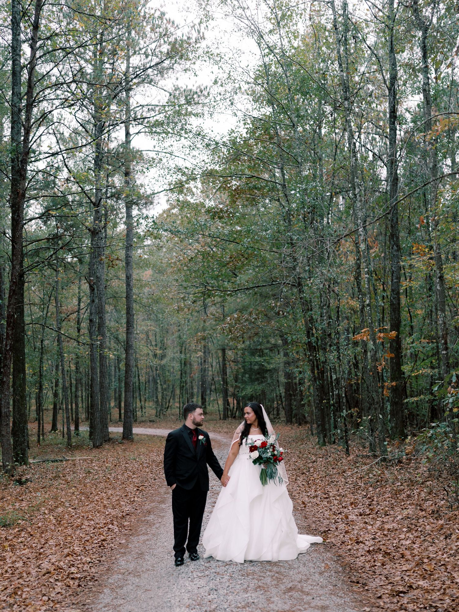 Wedding couple walks through autumn forest path surrounded by fallen leaves.