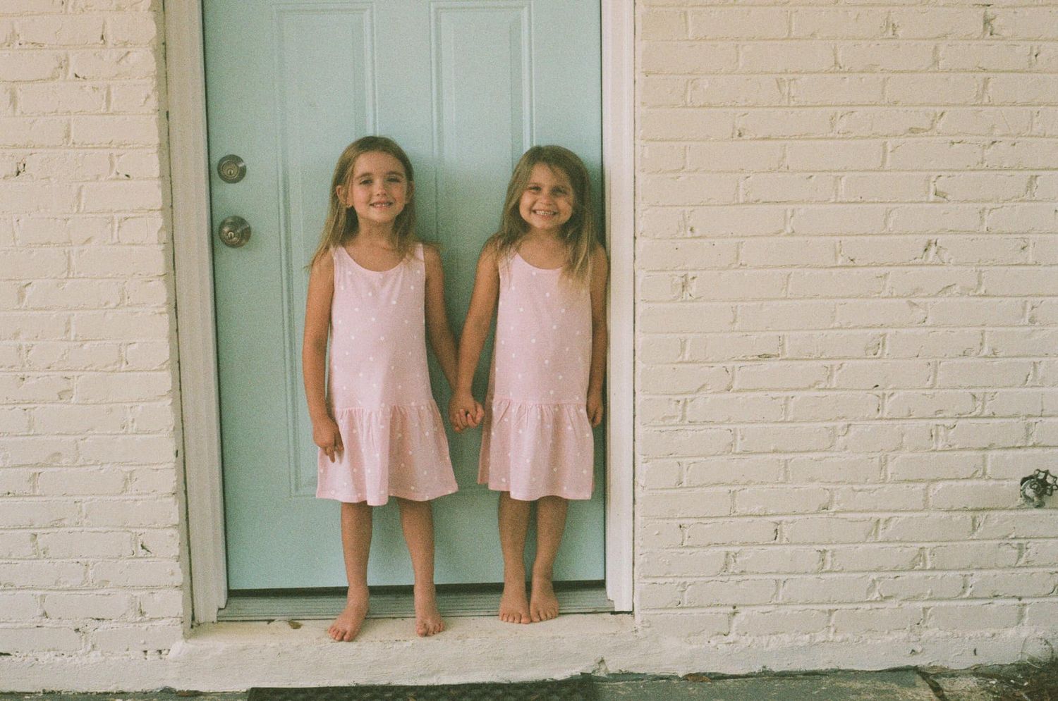 Two children in matching pink dresses standing in front of a mint green door.
