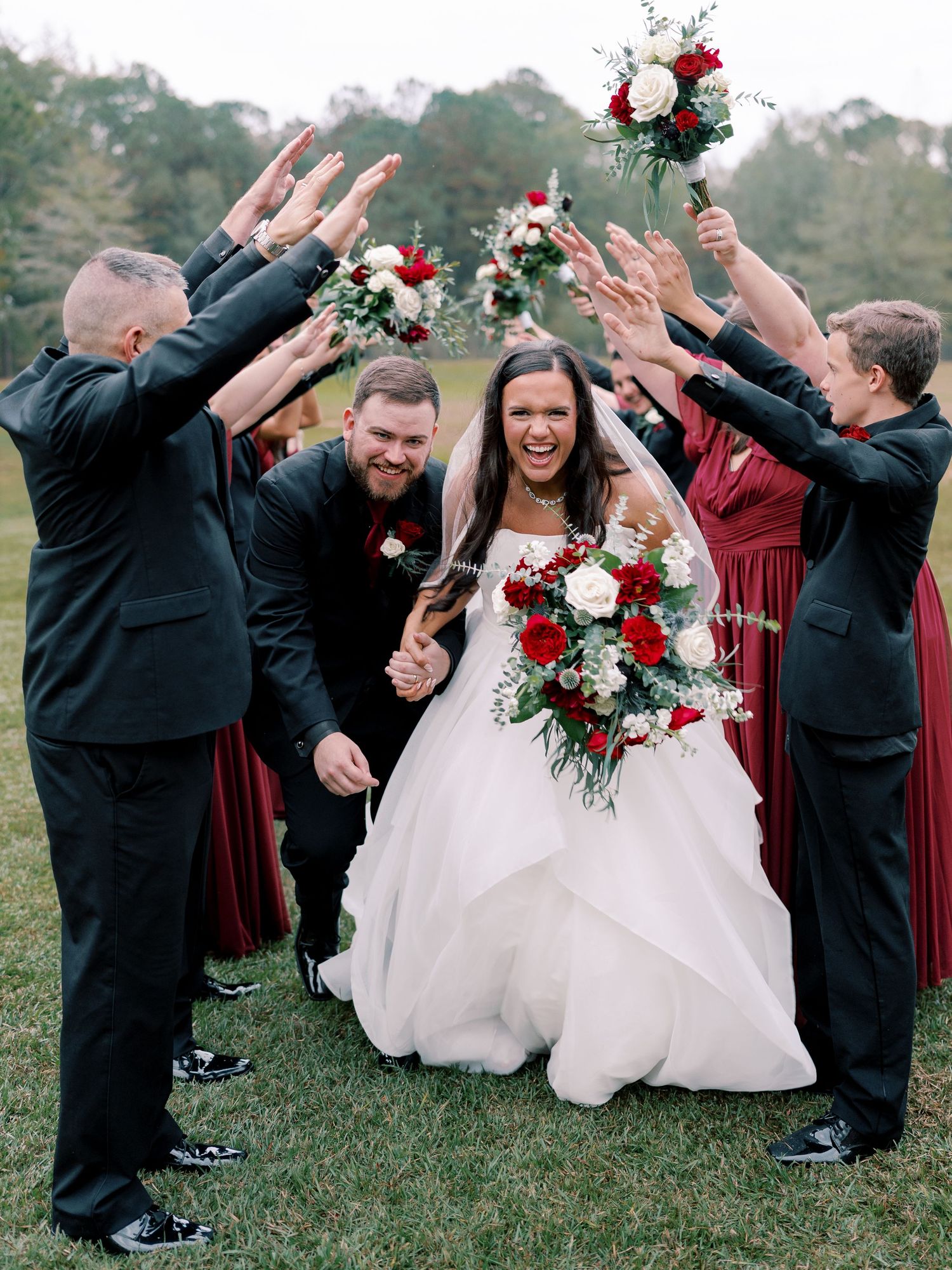Wedding party celebrates joyfully outdoors with red and white floral bouquets and burgundy bridesmaid dresses.