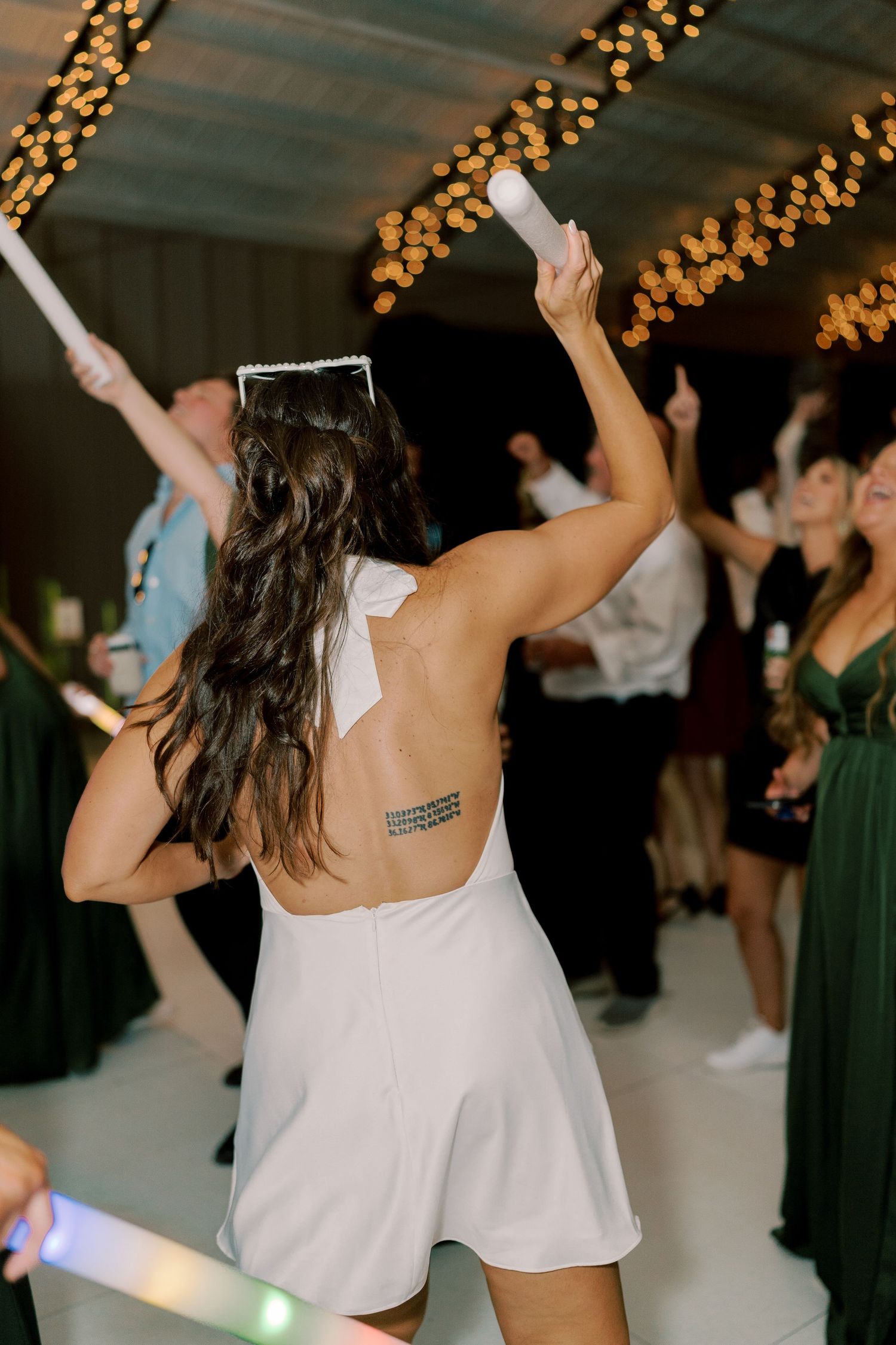 Dancing guest in white dress holding glow sticks under string lights at reception.
