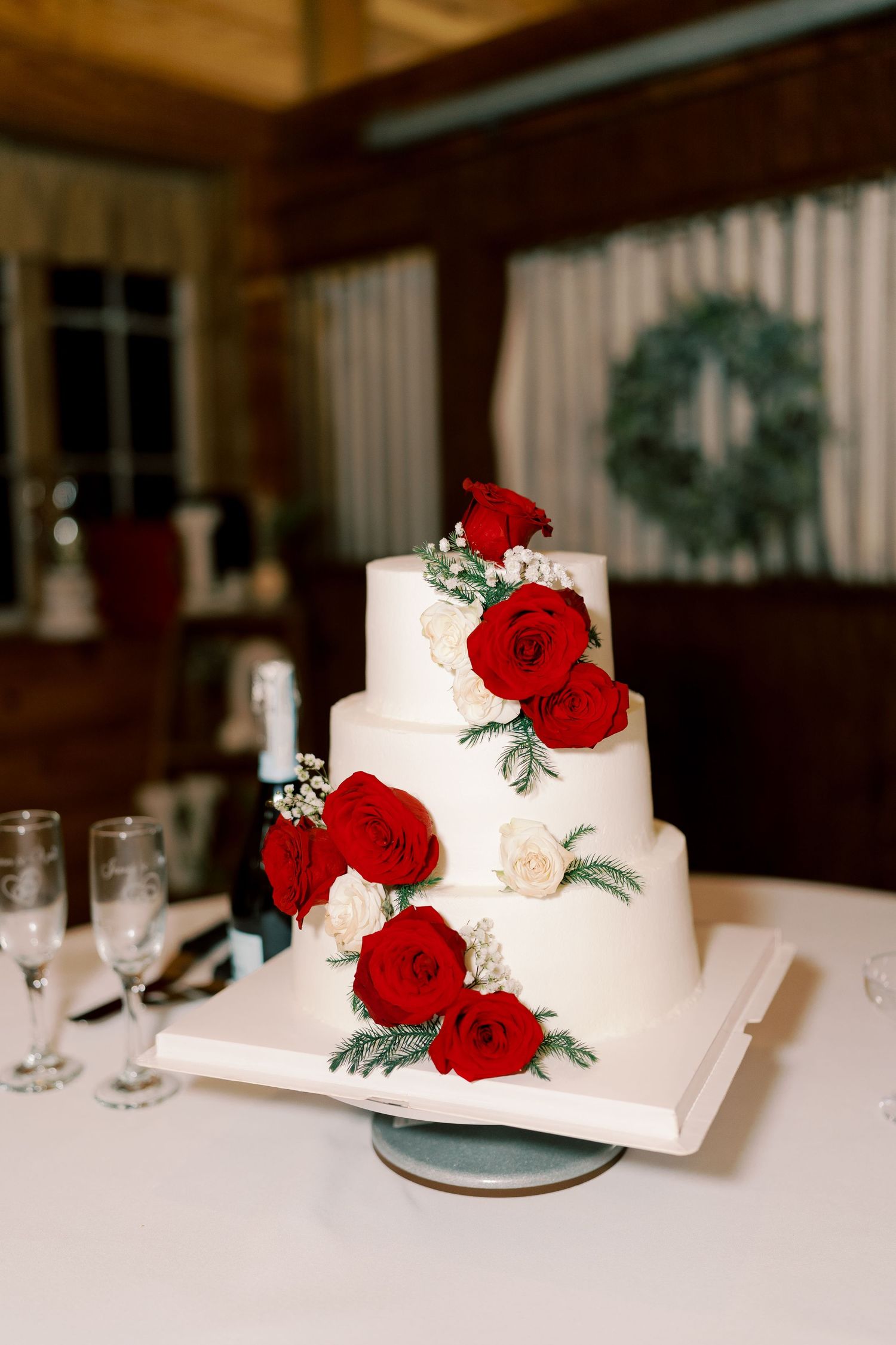 Elegant white wedding cake decorated with red roses and greenery on a white tablecloth.