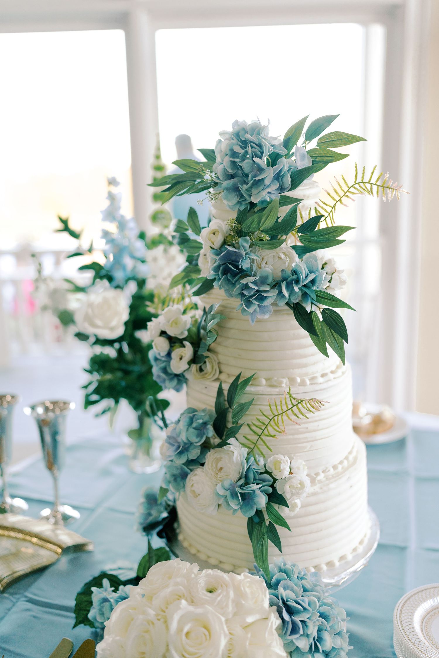 White tiered wedding cake decorated with blue hydrangeas and greenery on an elegant table setting.