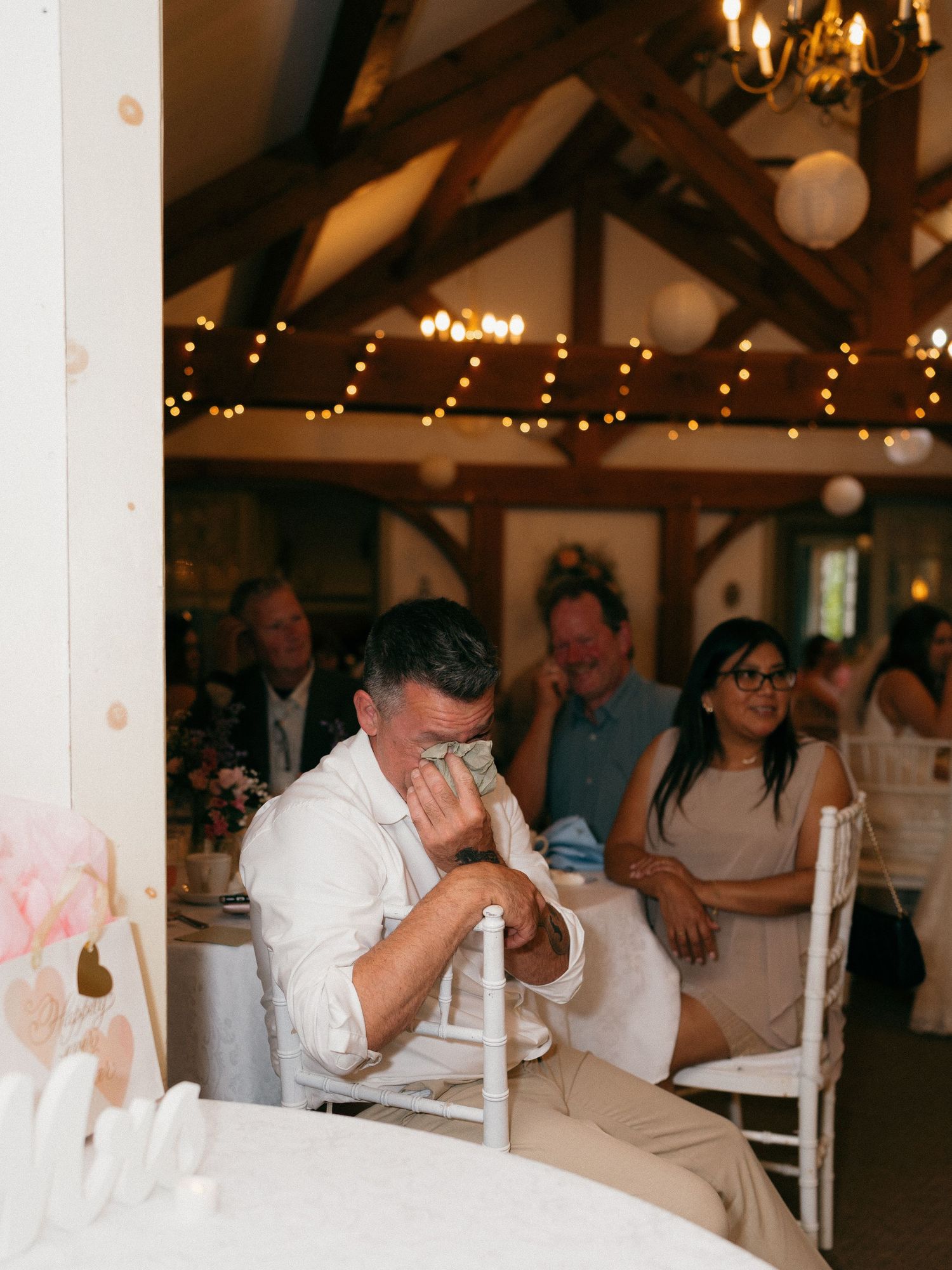 Wedding guests seated at tables in a rustic barn venue with exposed beams and string lights.