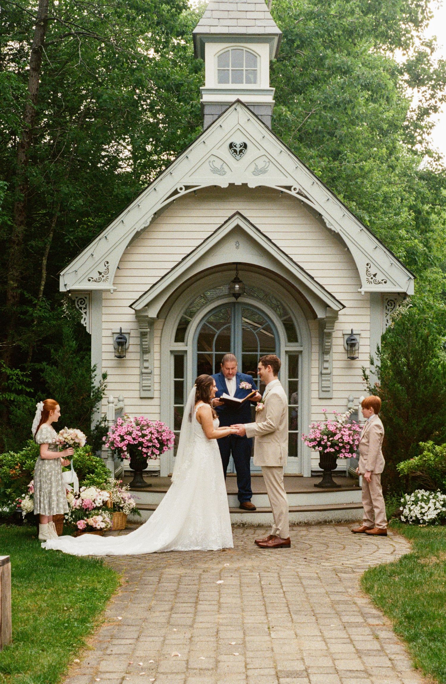 A wedding ceremony takes place outside a charming white chapel with pink floral arrangements and a stone pathway.