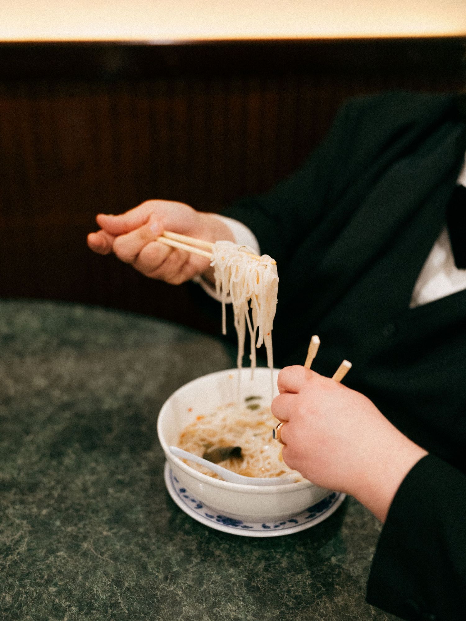 Close-up of hands lifting noodles with chopsticks from a white bowl.
