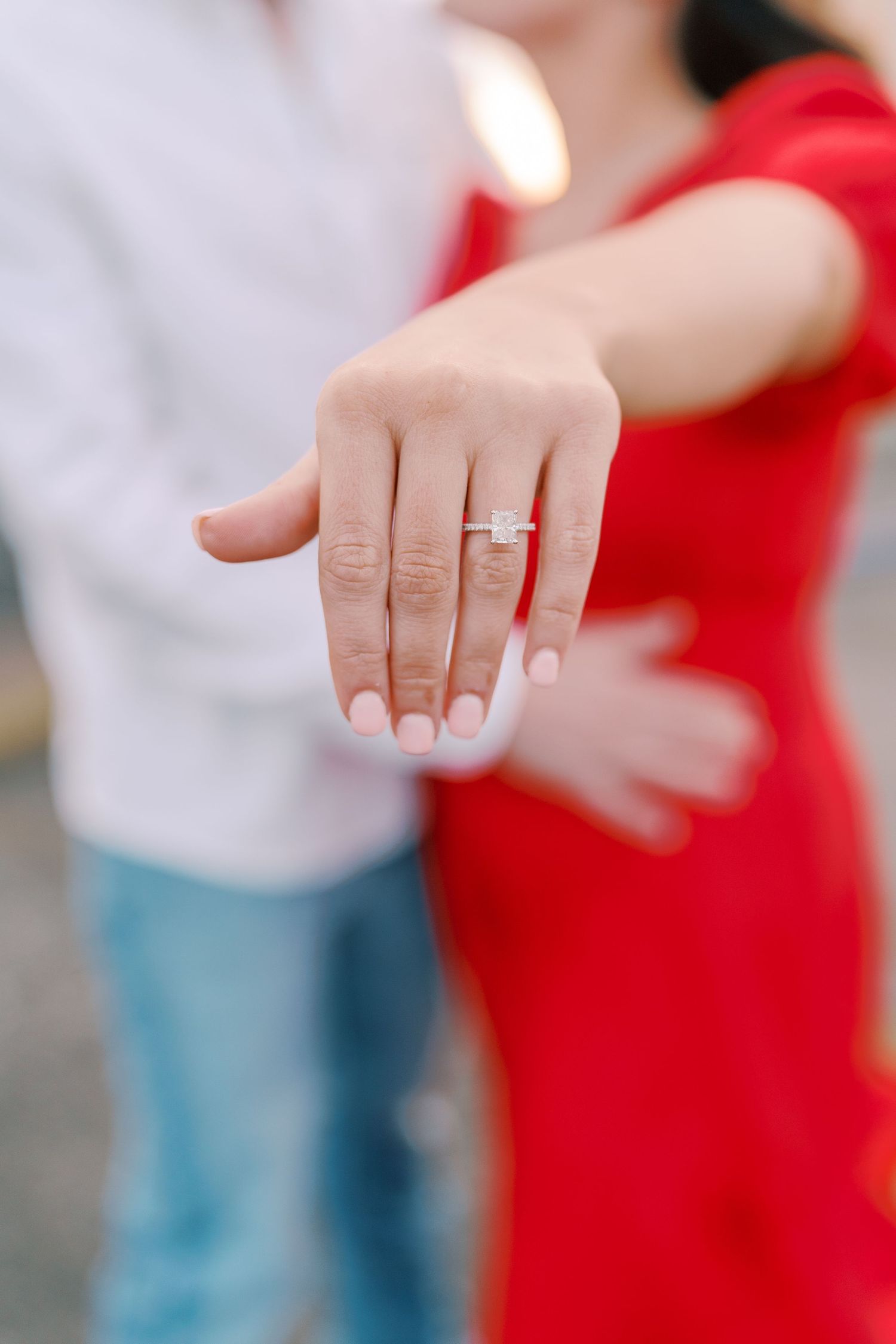Close-up of hands showing off an engagement ring against blurred red and white clothing.