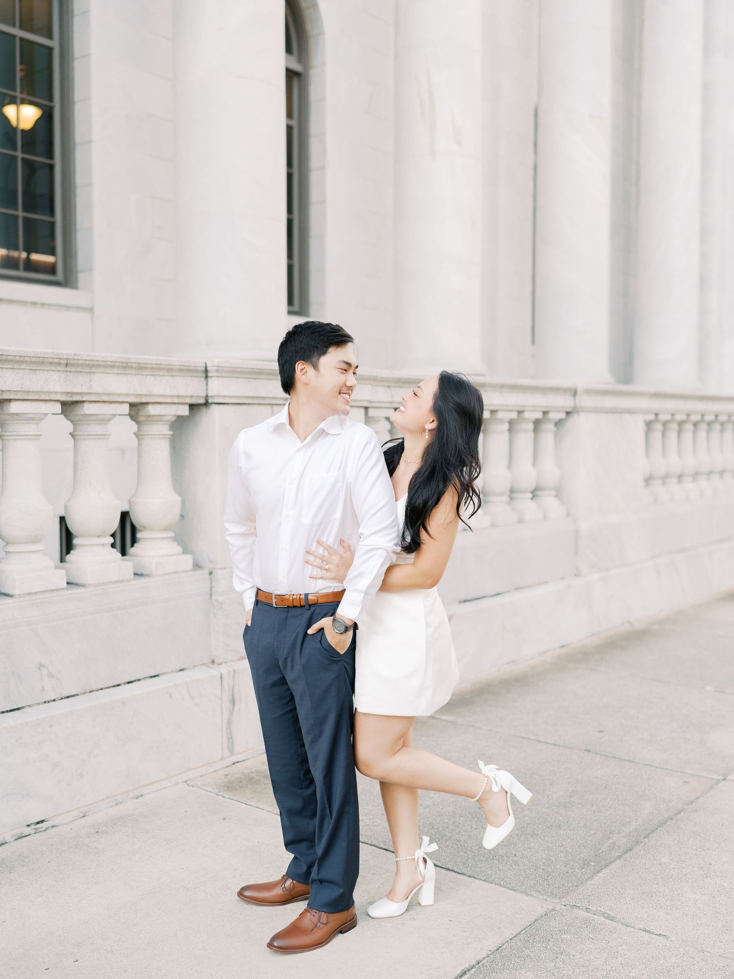 A couple shares playful poses along a classical white columned building exterior.