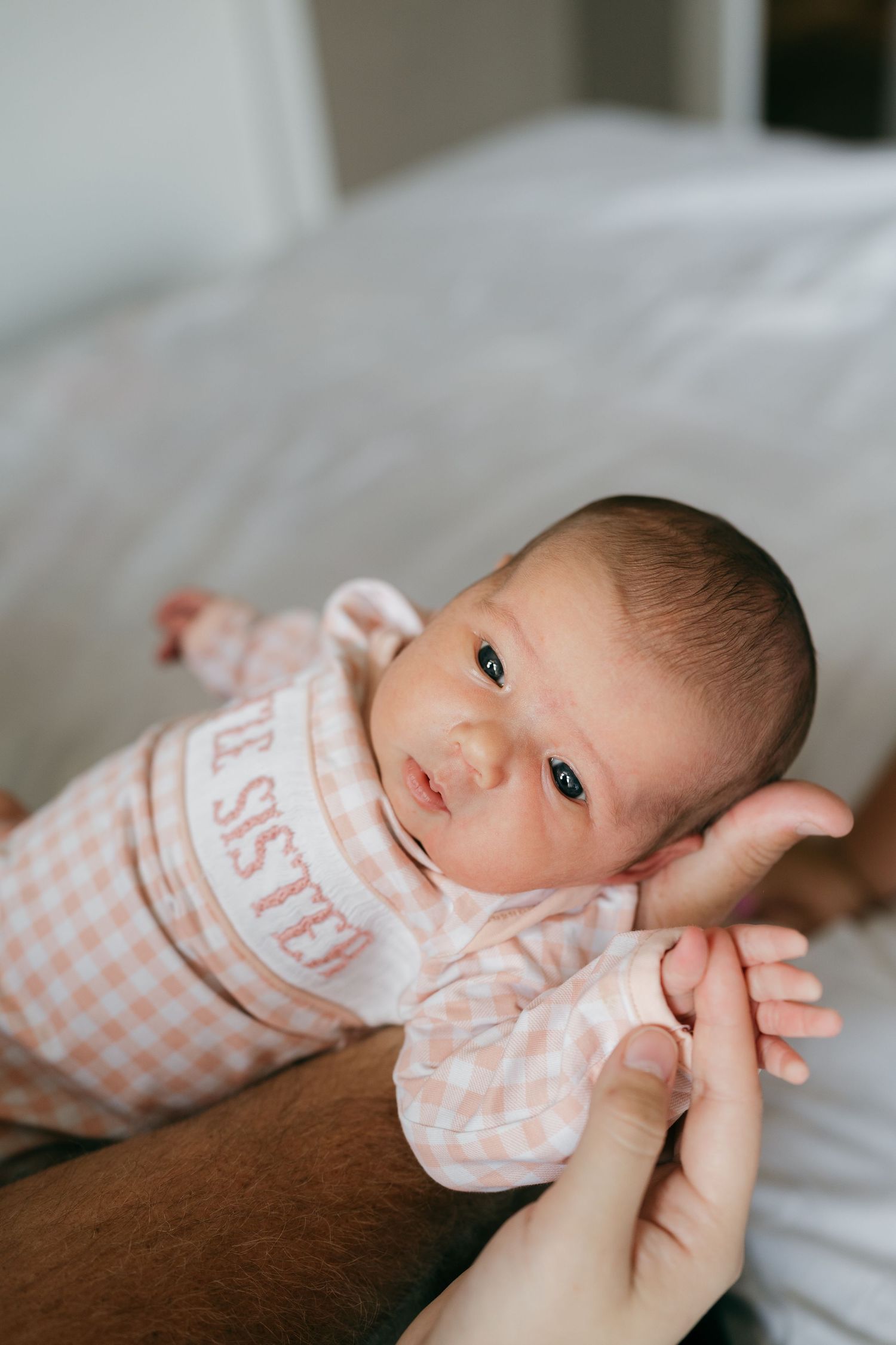 Baby in pink checkered outfit lying on white bedding during portrait session.