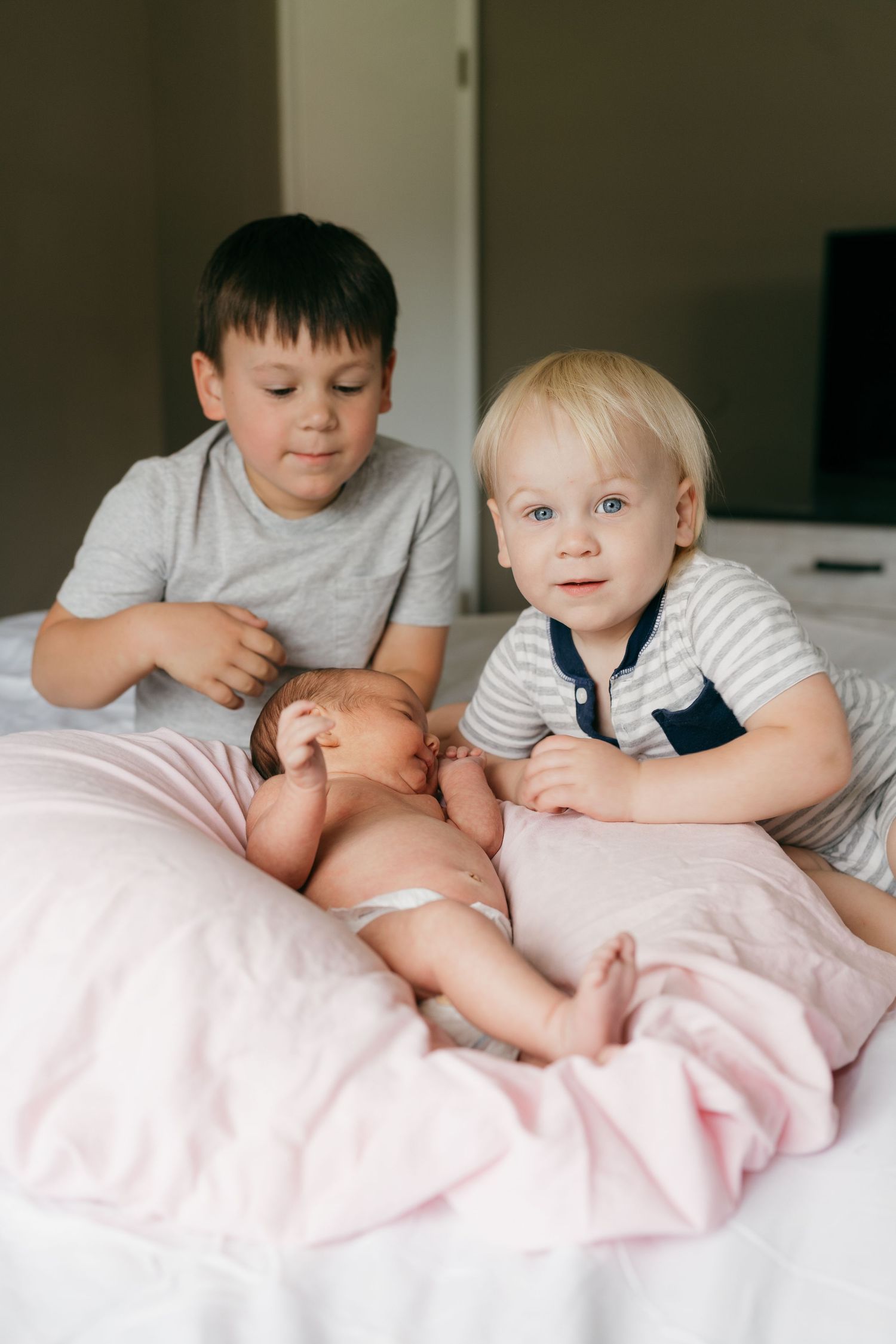 Children sharing sweet moments with baby sibling on bed with soft pink bedding.